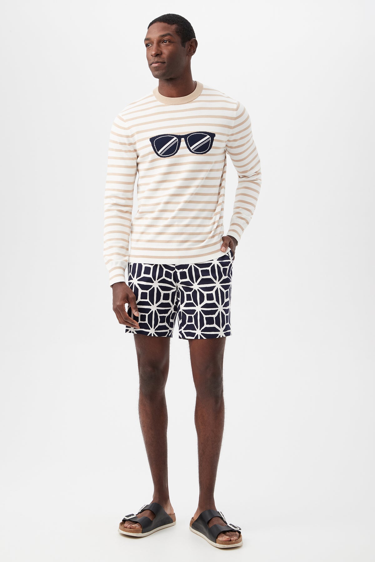 A man stands against a white background wearing a cream and beige striped sweater with a sunglasses graphic, Lawrence Short - Durell Herringbone, and black sandals.