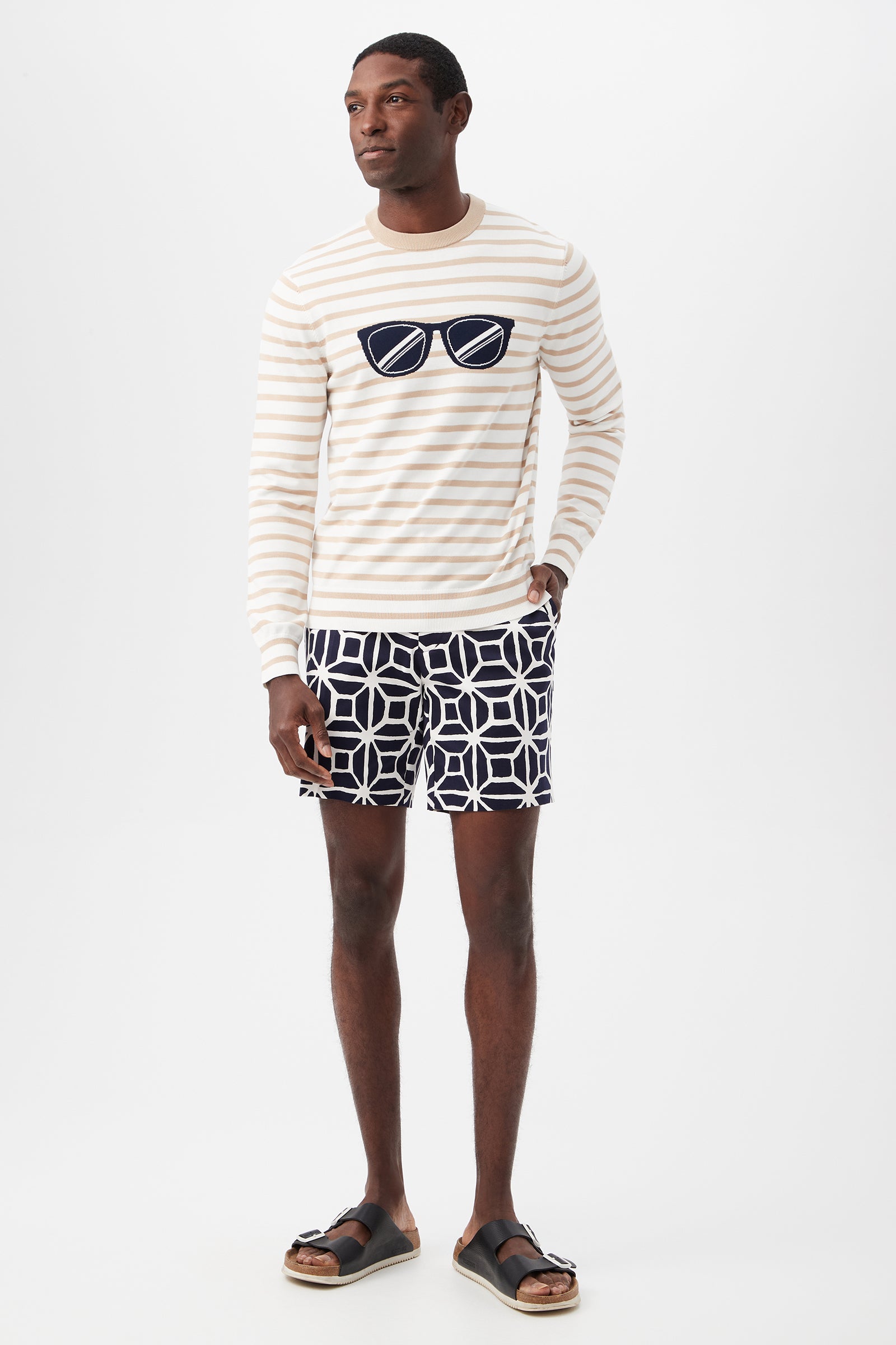 A man stands against a white background wearing a cream and beige striped sweater with a sunglasses graphic, Lawrence Short - Durell Herringbone, and black sandals.