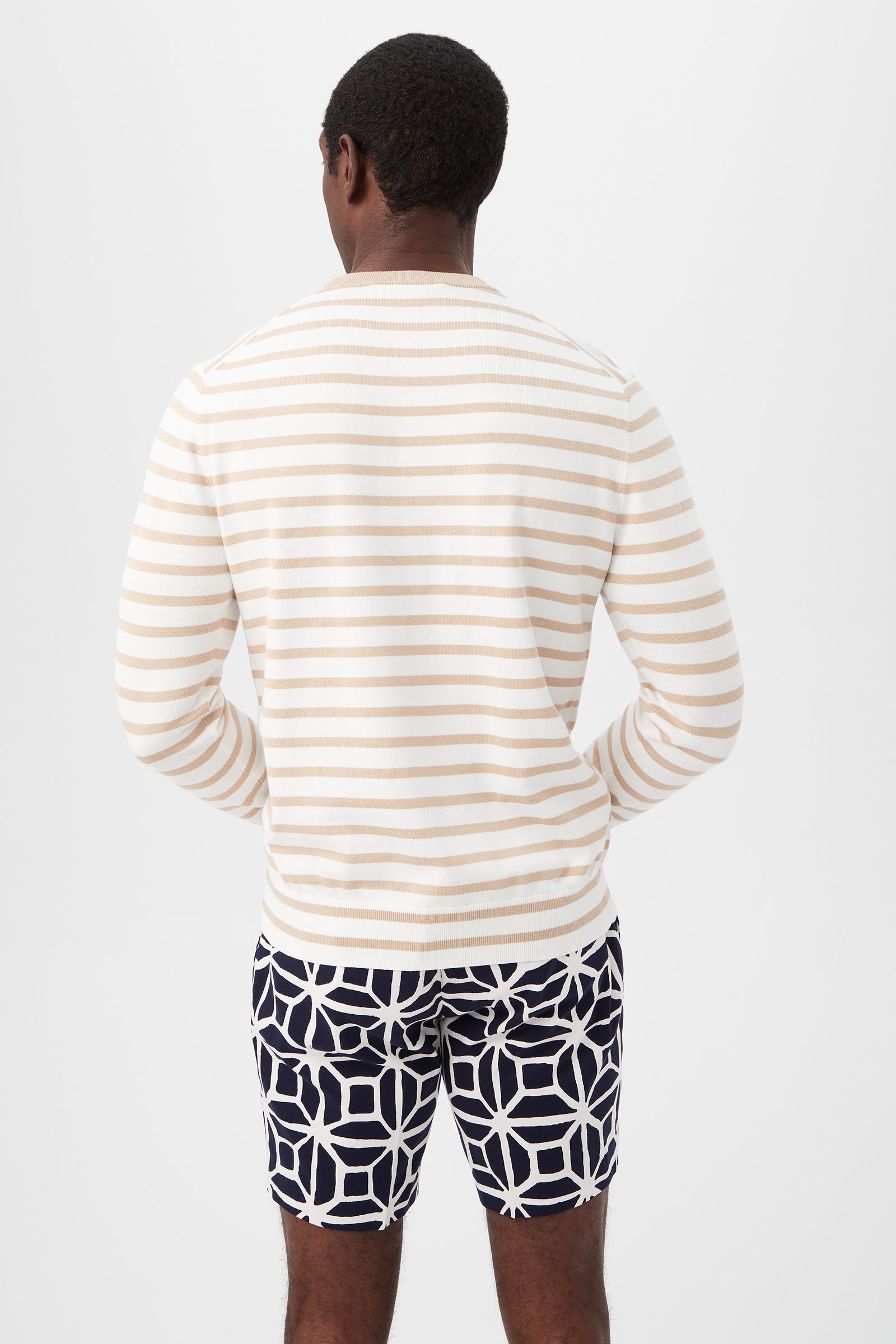 A man stands with his back to the camera, wearing the Julian Sunglasses Crewneck Sweater, a long-sleeve cream and beige striped top, paired with navy blue shorts featuring a bold white geometric pattern, set against a plain white background.