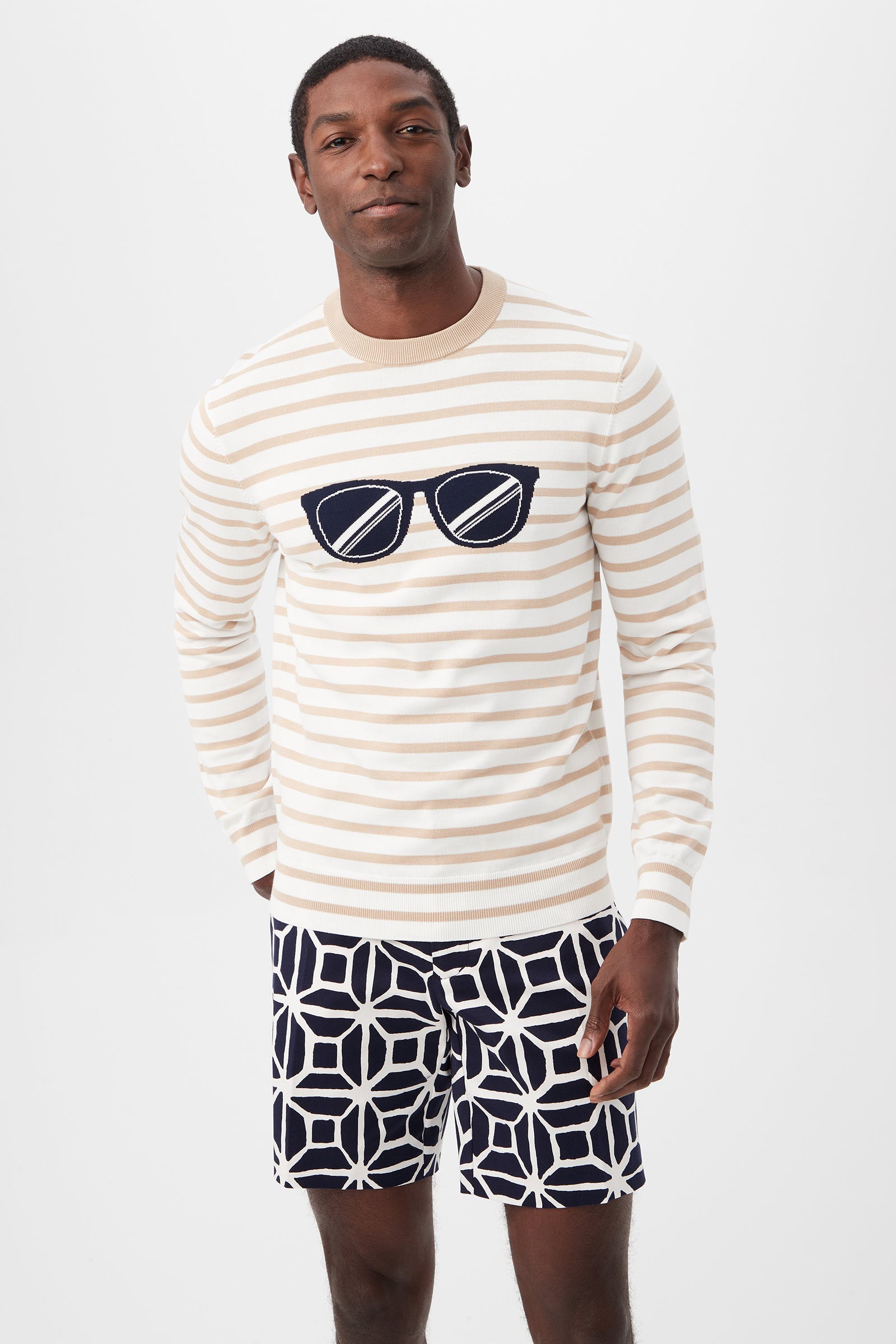 A man stands against a plain white background wearing the Julian Sunglasses Crewneck Sweater in Optic White Multi and navy and white geometric patterned shorts.