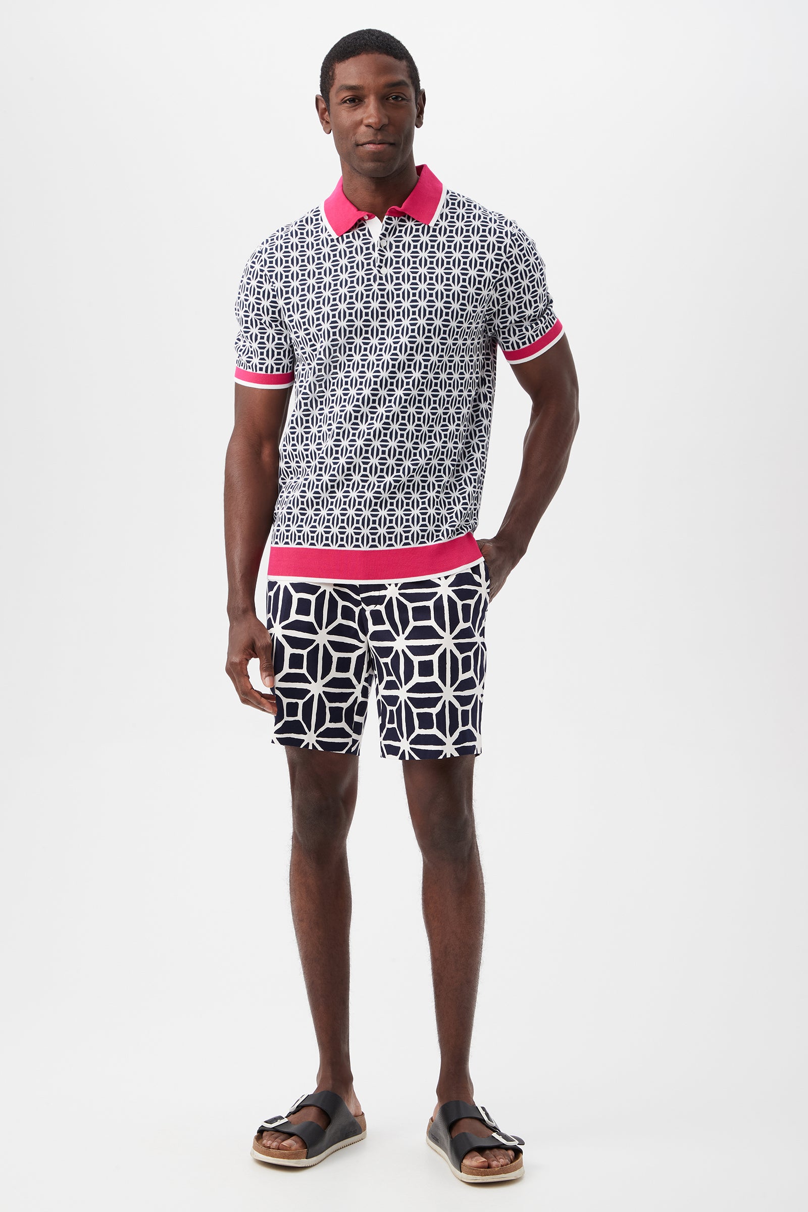 A man stands against a white background wearing the Grayson Short-Sleeve Polo with a pink collar and trim, navy and white geometric shorts, and black sandals.