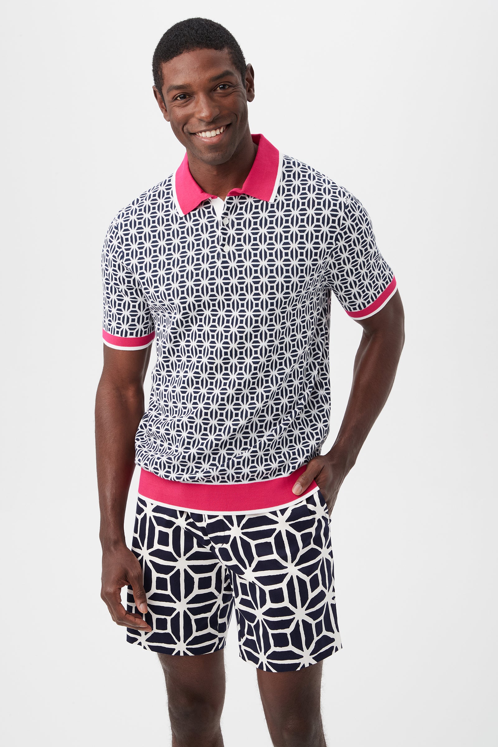 A smiling man wears the Grayson Short-Sleeve Polo in Indigo Multi, featuring a navy and white geometric pattern with a bright pink collar and cuffs, paired with matching shorts, standing against a plain white background.