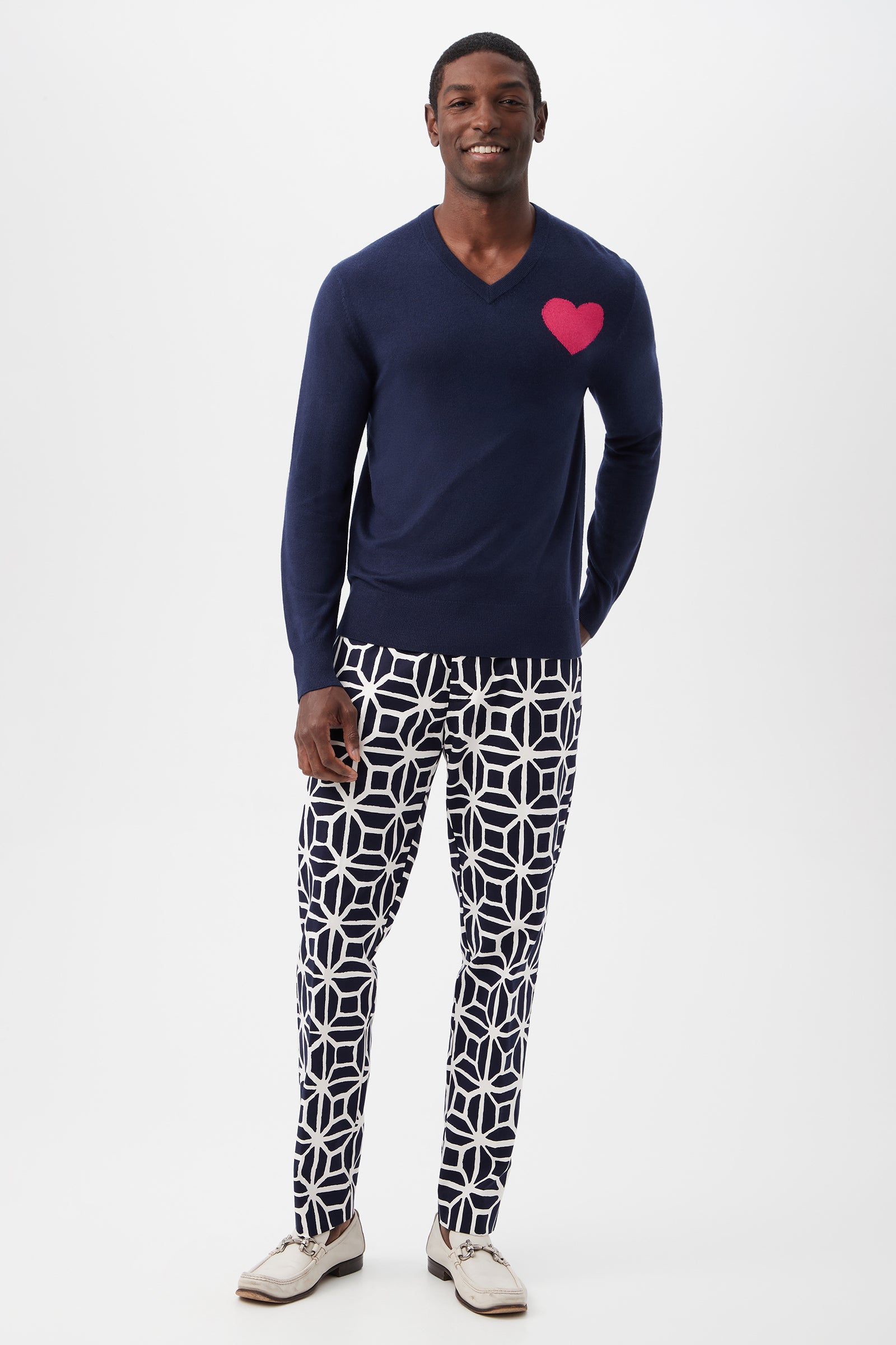 A man smiles in a Lover Heart V-Neck Sweater, featuring navy blue fabric and a pink chest heart, paired with black-and-white geometric pants and white dress shoes. The background is plain white.