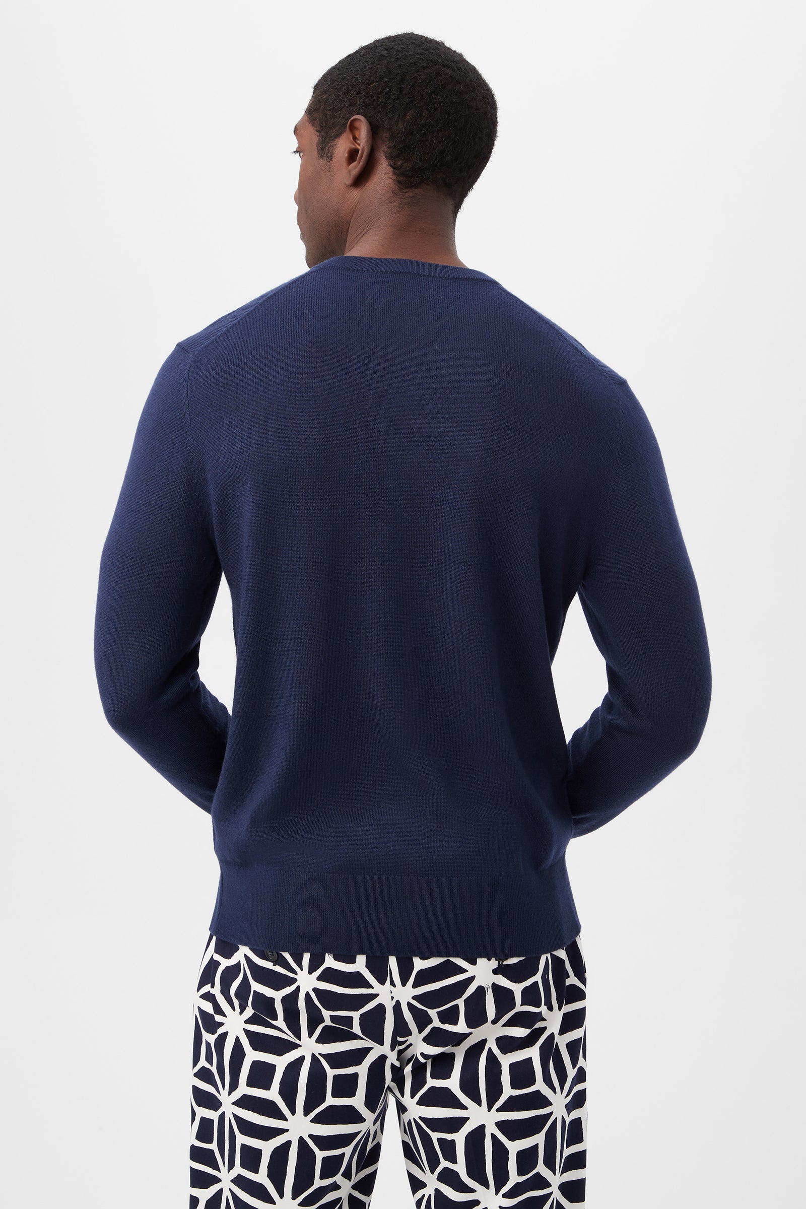 A man stands with his back to the camera, wearing the Lover Heart V-Neck Sweater in dark blue and white pants with a bold geometric navy pattern, set against a plain white background.