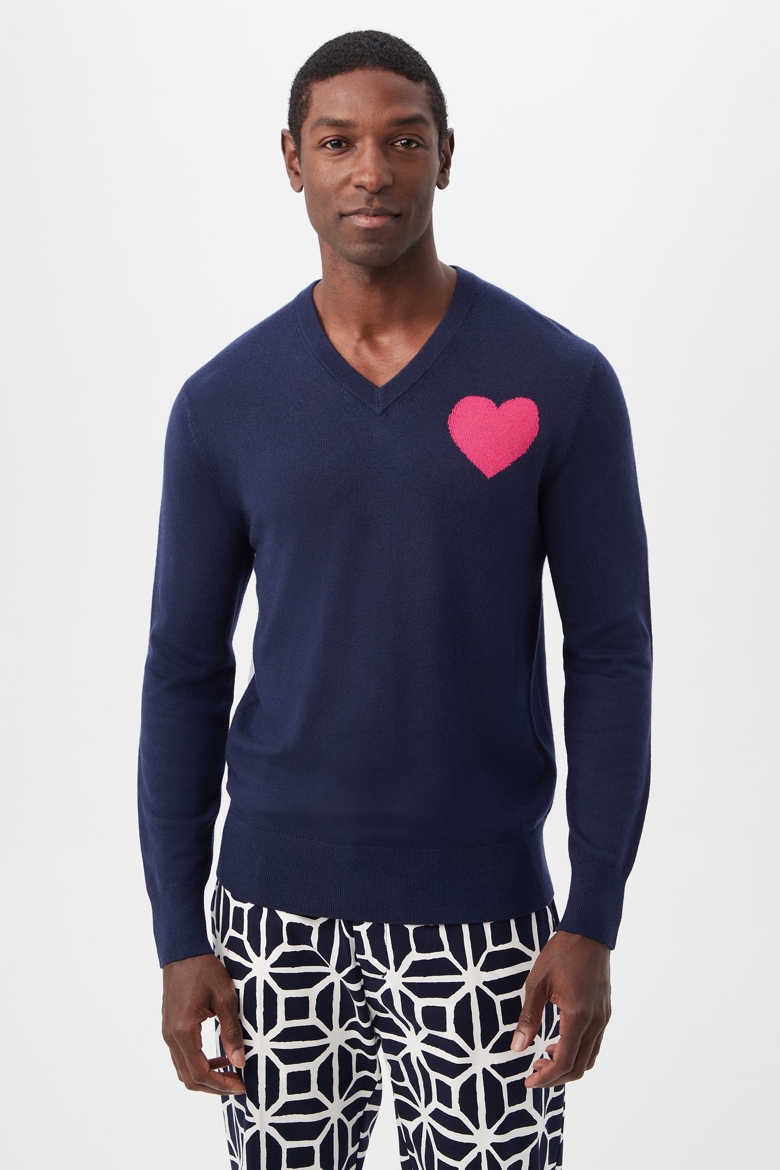 A man wearing the Lover Heart V-Neck Sweater in Indigo/Petal Pink with a heart on the left chest and geometric navy-and-white pants stands against a plain white background.