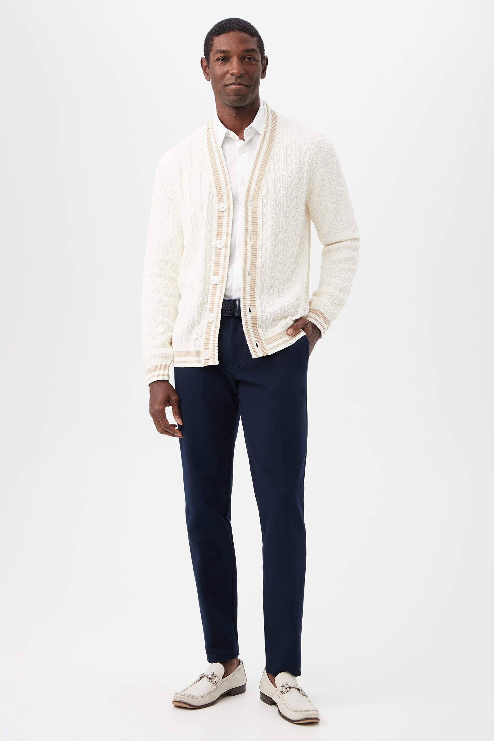 A man stands against a white background wearing the Lucien Cotton V-Neck Cardigan over a white shirt, paired with navy trousers and light loafers. One hand is in his pocket as he smiles slightly.