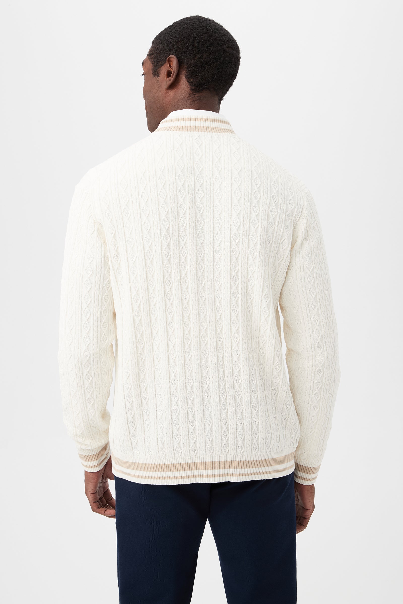 A man is seen from behind wearing the Lucien Cotton V-Neck Cardigan in cream, featuring cable-knit details and ribbed cuffs and collar, paired with dark navy pants against a plain white background.