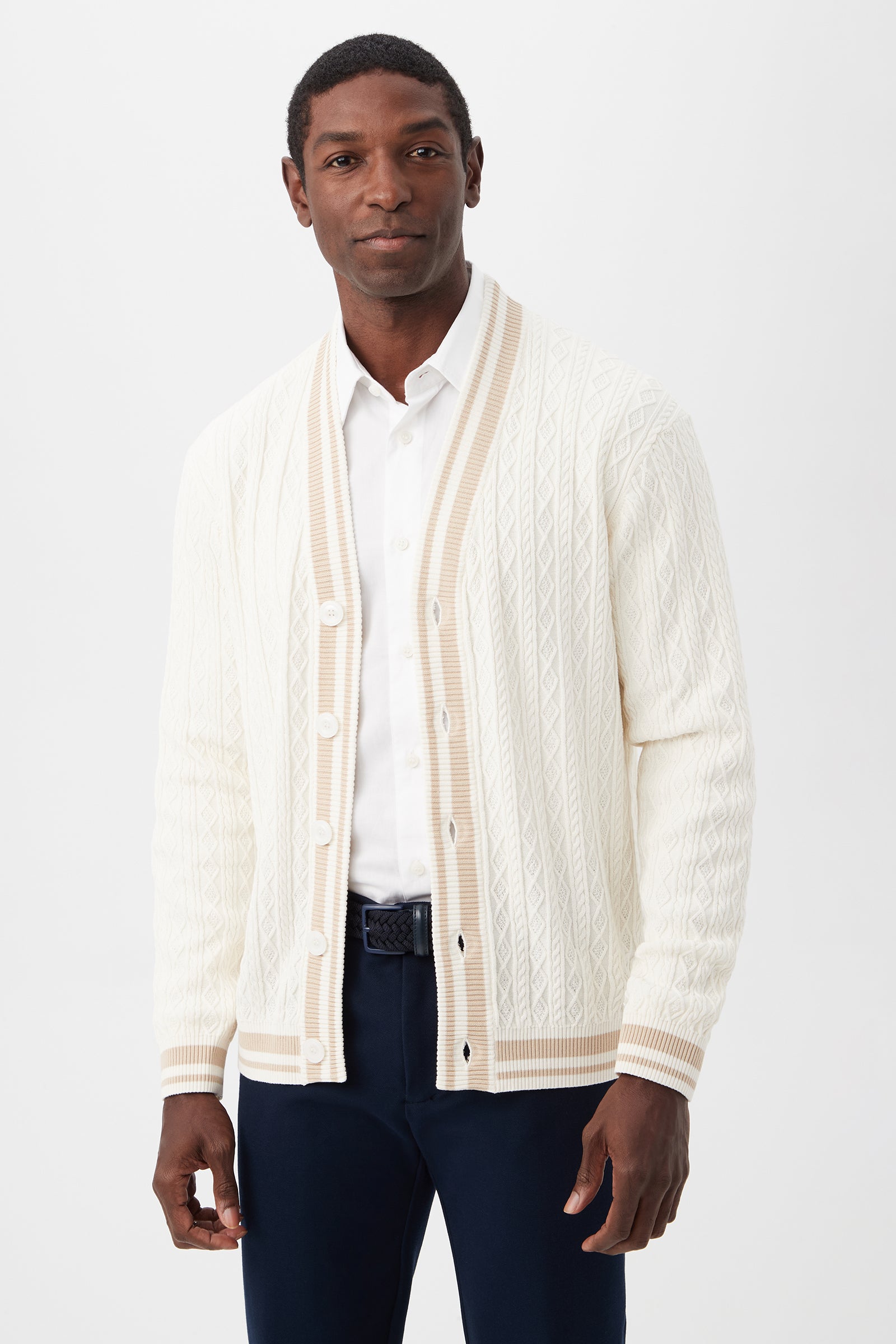 A man stands against a plain background wearing the Lucien Cotton V-Neck Cardigan in Optic White/Chai over a white collared shirt and dark trousers.