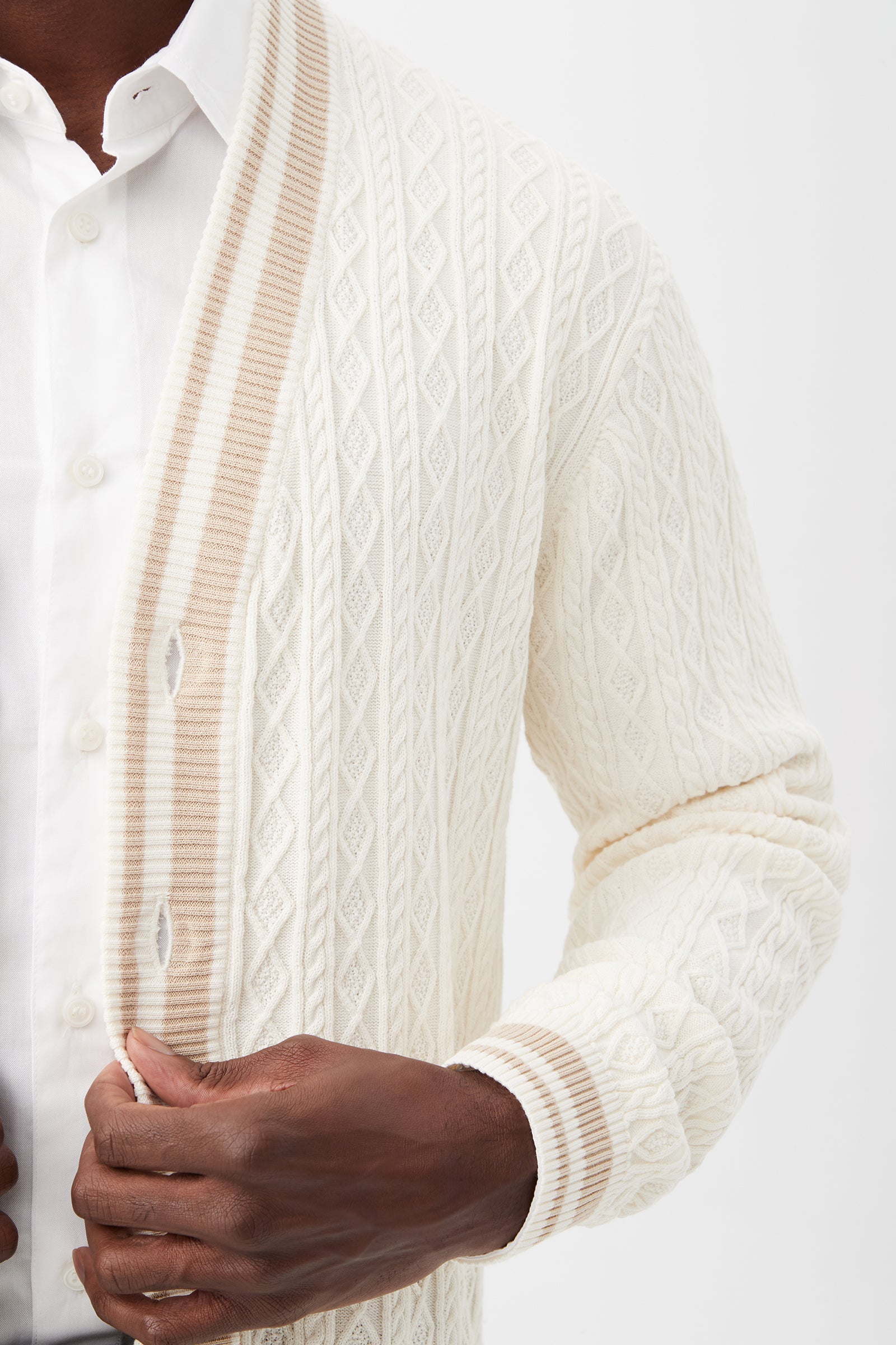 A person adjusts the Lucien Cotton V-Neck Cardigan, an off-white cable-knit cardigan with beige striped trim, layered over a white button-up shirt and shown from shoulders to waist.
