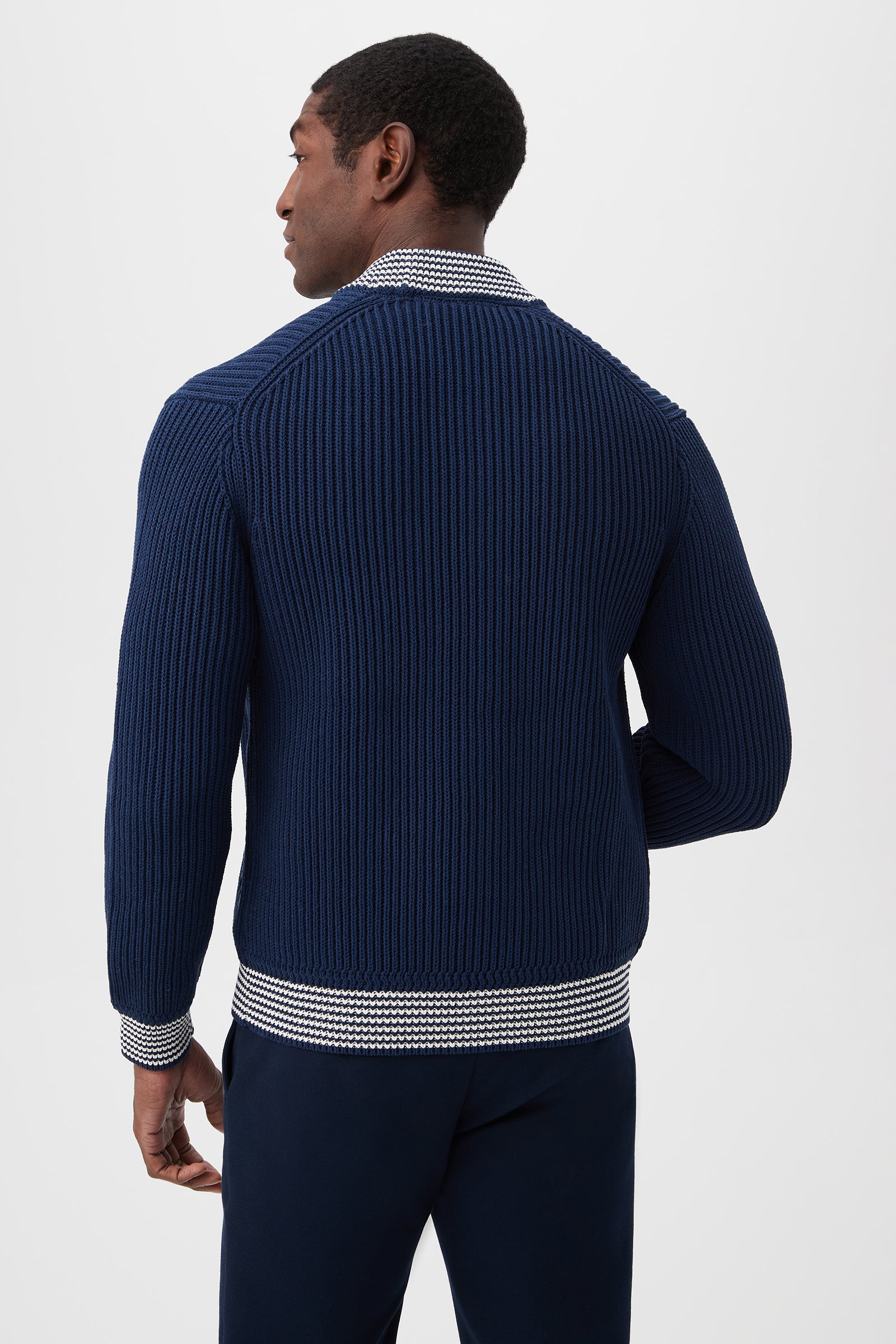 A man stands facing away, wearing the Robertson Bomber Jacket - Magic Cotton in navy blue with patterned white and navy trim on the collar, cuffs, and hem, paired with dark trousers against a plain light background.