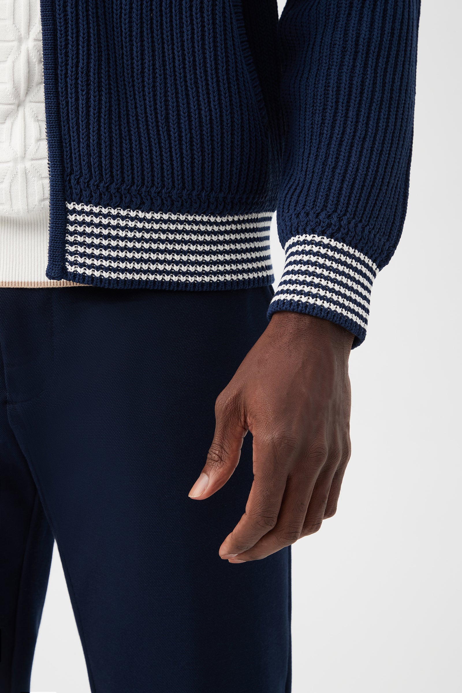 A close-up of a person wearing the Robertson Bomber Jacket in navy Magic Cotton, featuring ribbed texture and white striped cuffs, layered over a white textured shirt and paired with navy pants. Their left arm rests relaxed by their side.