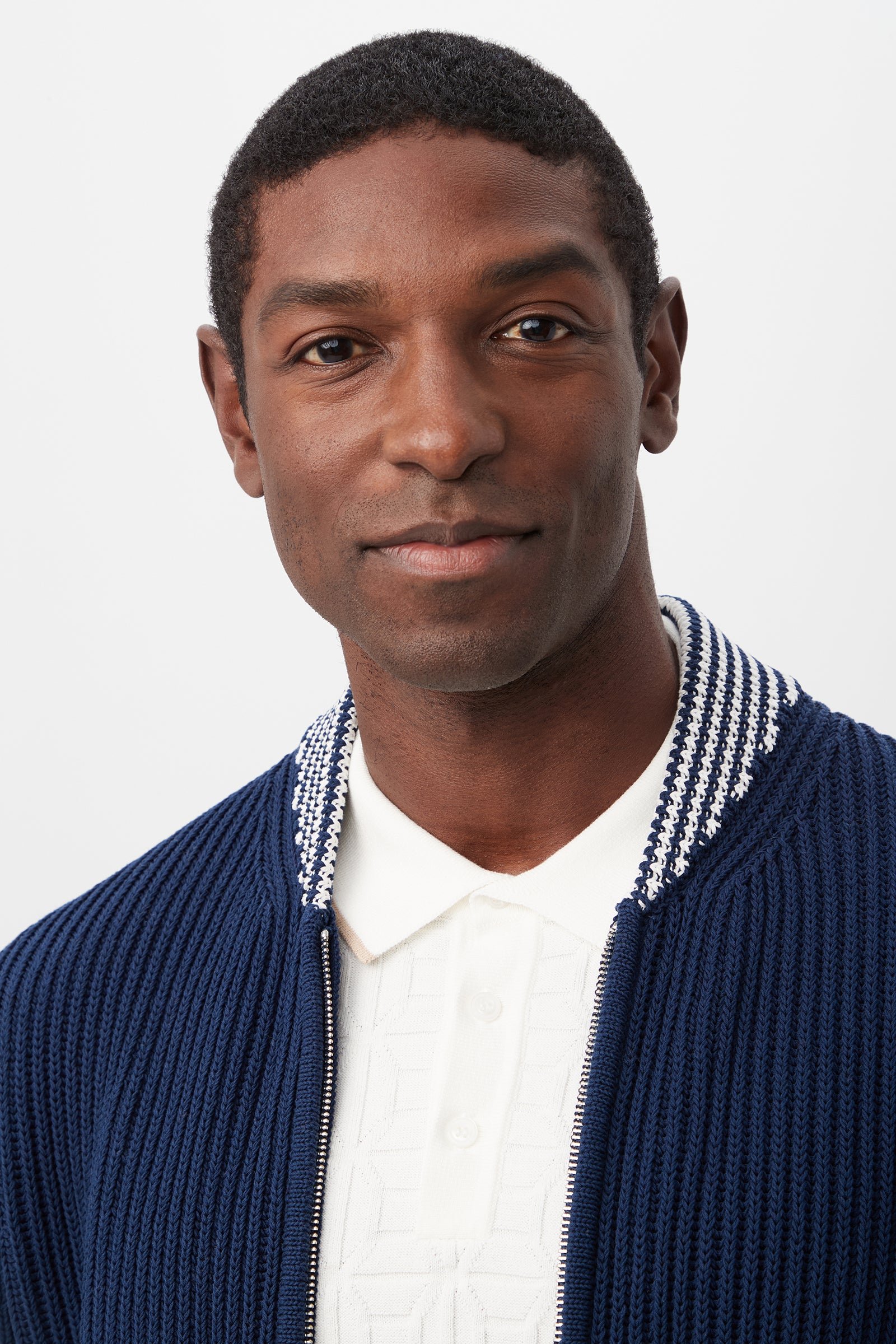 A man with short curly hair wears a white collared shirt and the Robertson Bomber Jacket - Magic Cotton, which features a patterned collar. He looks at the camera and smiles slightly against a plain light background.