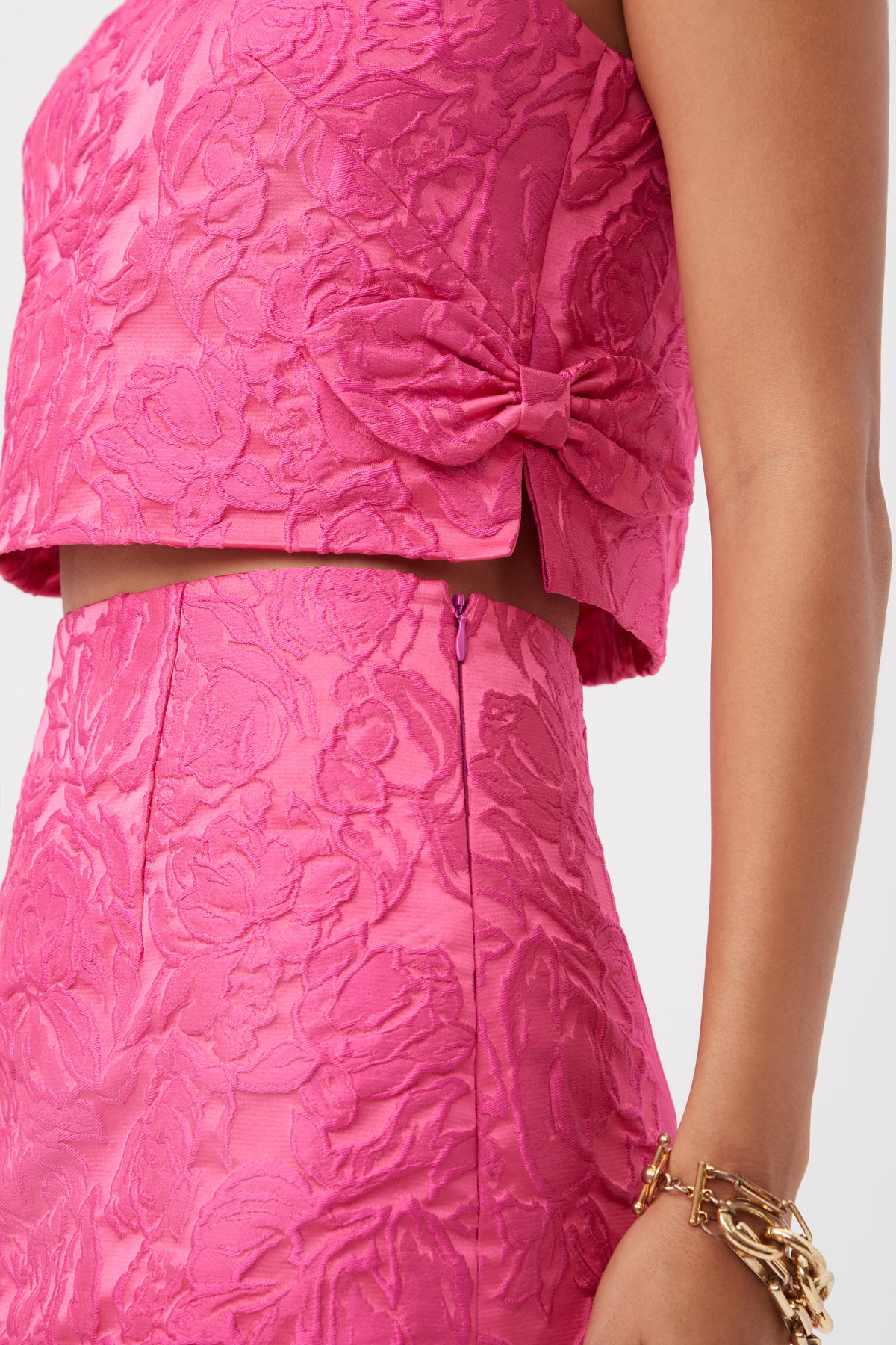 A person wears the Cayley Top - Sweetheart Jacquard, a pink floral-textured sleeveless top with a side bow, paired with a matching skirt and a gold bracelet.