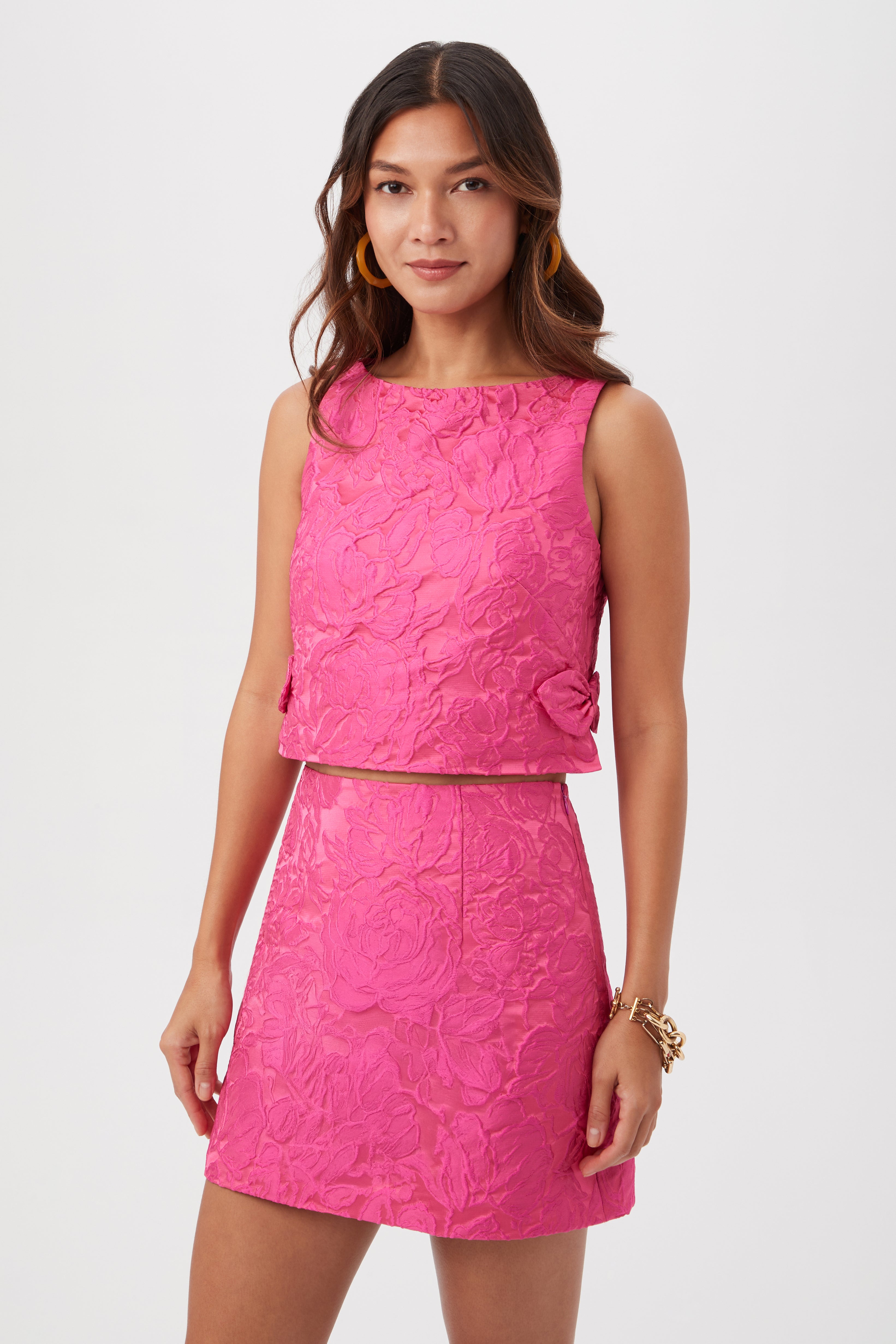 A woman wears the High Rise Rico Skirt - Sweetheart Jacquard, a sleeveless bright pink textured crop top, and matching mini skirt with floral patterns. She stands against a white background with long wavy hair, gold hoop earrings, and a bracelet.