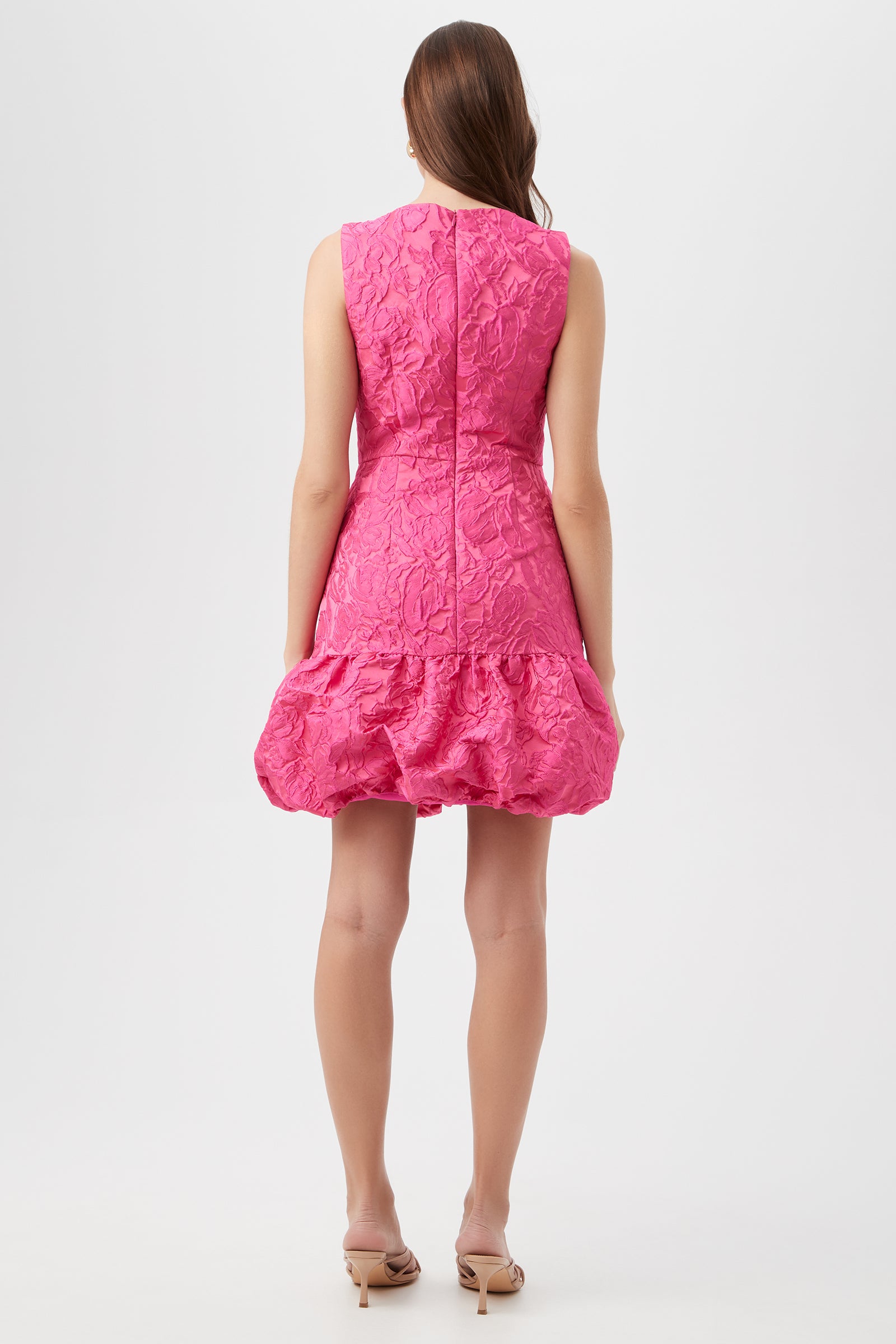 A woman with long brown hair stands facing away, wearing the Chiara Dress - Sweetheart Jacquard, a sleeveless textured pink dress with a ruffled hem, paired with nude heeled sandals against a plain light background.
