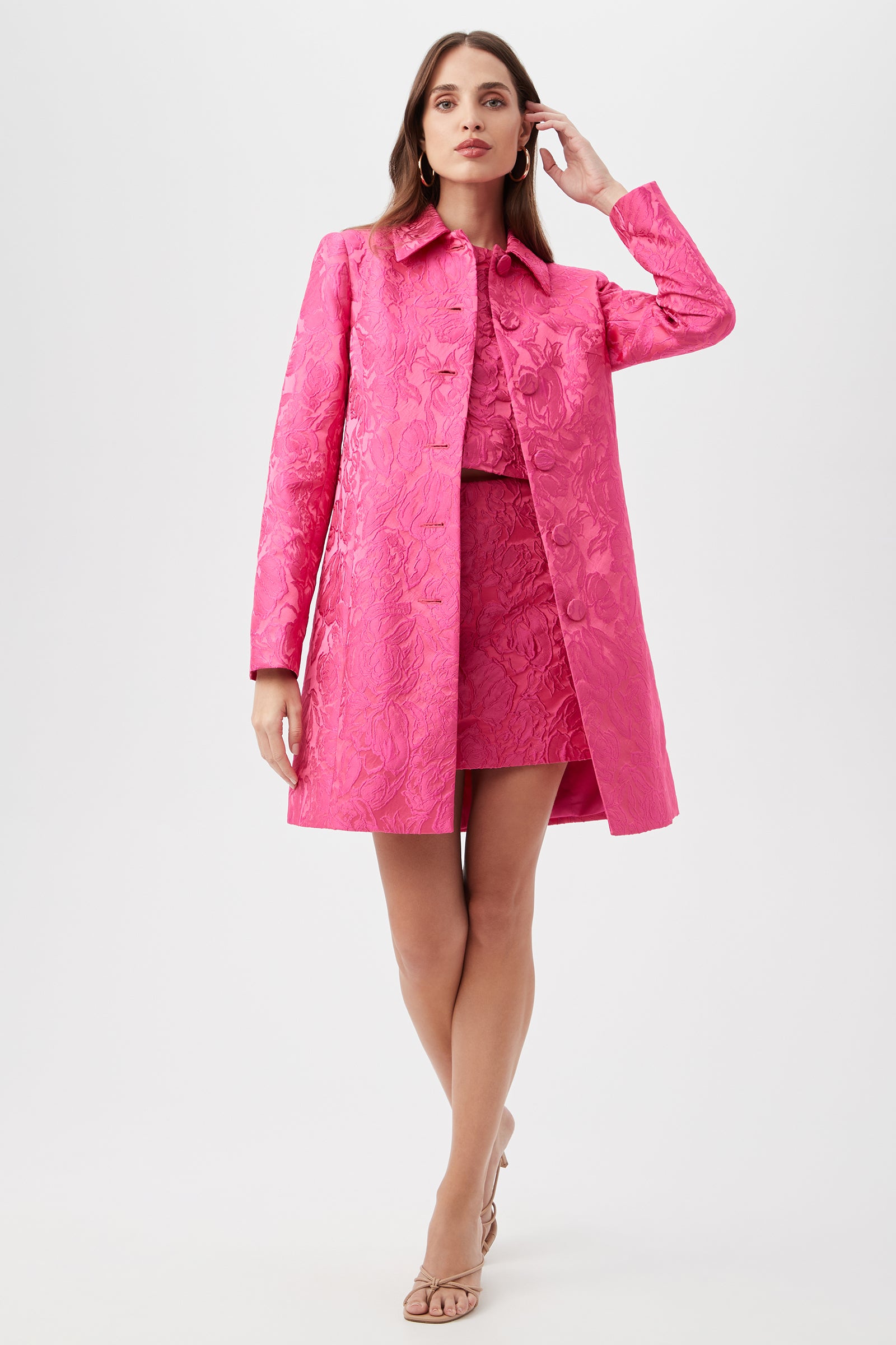 A woman poses in the Asher Coat - Sweetheart Jacquard in Snapdragon over a matching skirt and top, touching her hair and wearing nude heels against a plain white background.