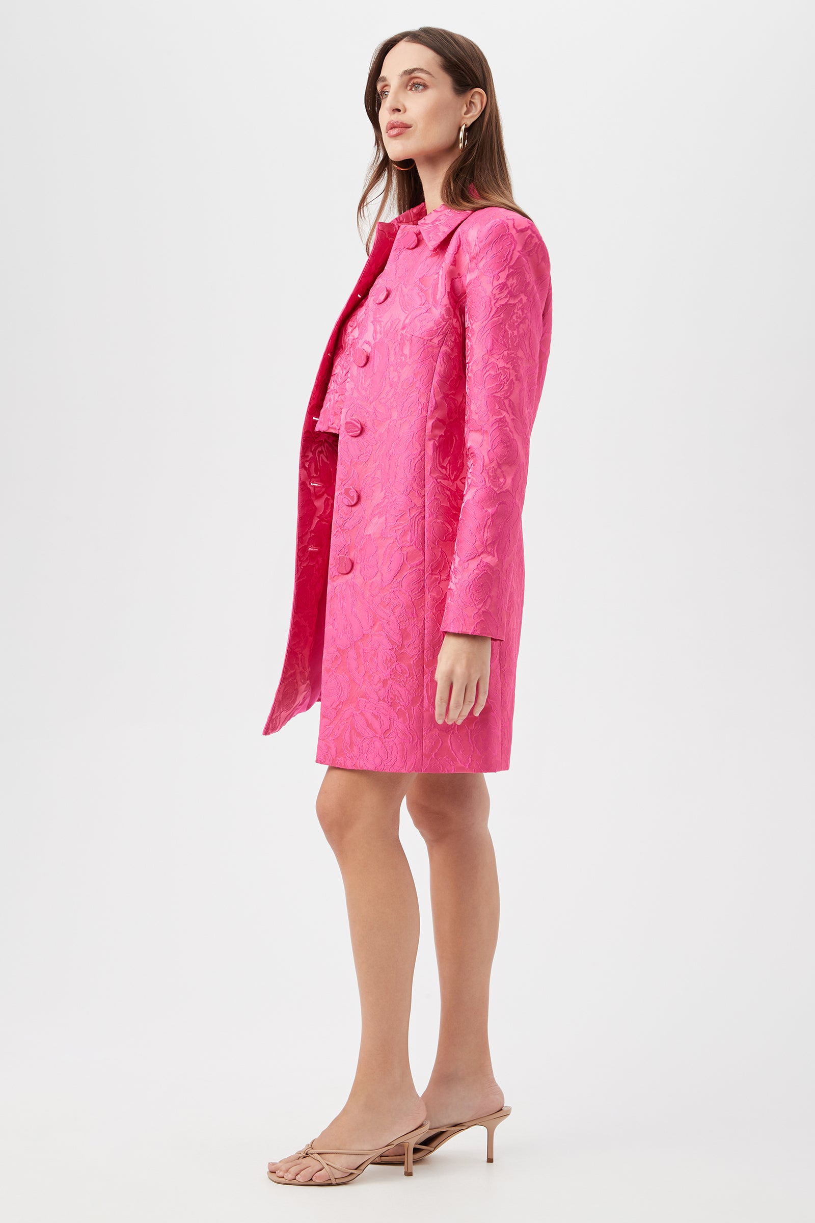 A woman stands sideways in the bright pink Asher Coat - Sweetheart Jacquard, knee-length with large buttons, paired with a matching skirt. She has long brown hair and wears nude high-heeled sandals against a plain white background.