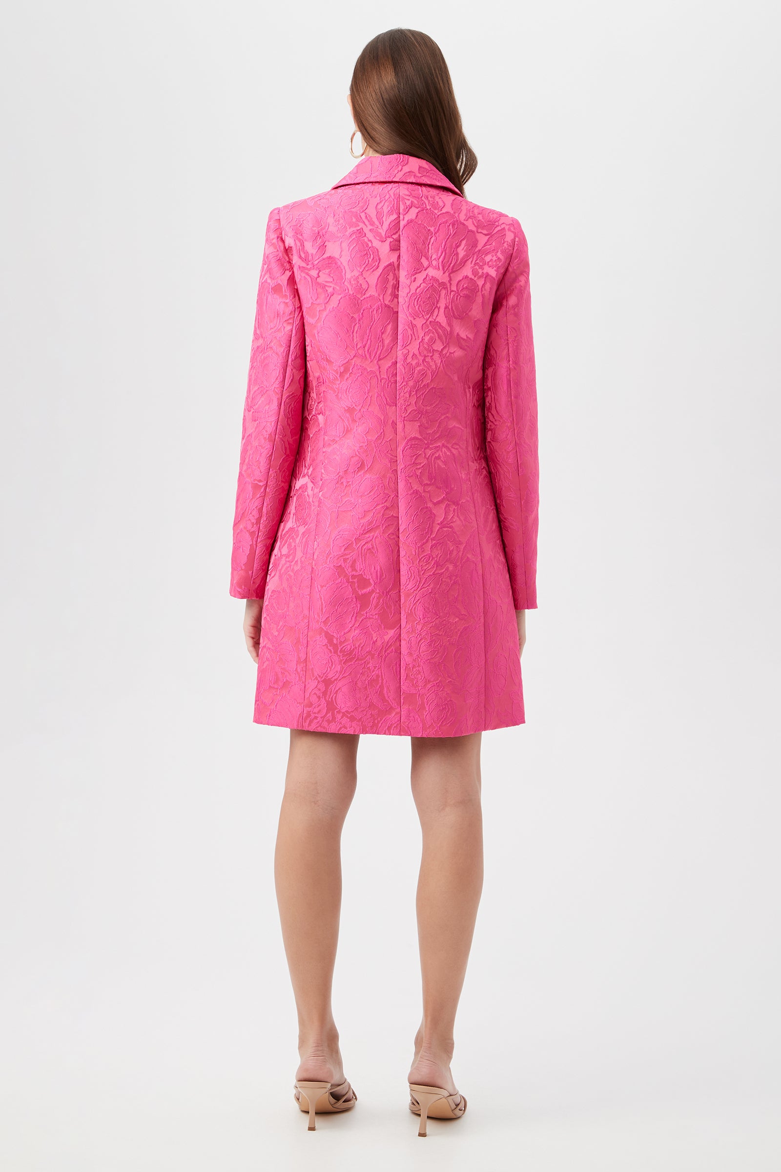 A woman with long brown hair stands facing away, wearing the Asher Coat - Sweetheart Jacquard: a bright pink, textured, long-sleeved coatdress with a floral pattern. She pairs it with beige heeled sandals against a plain white background.