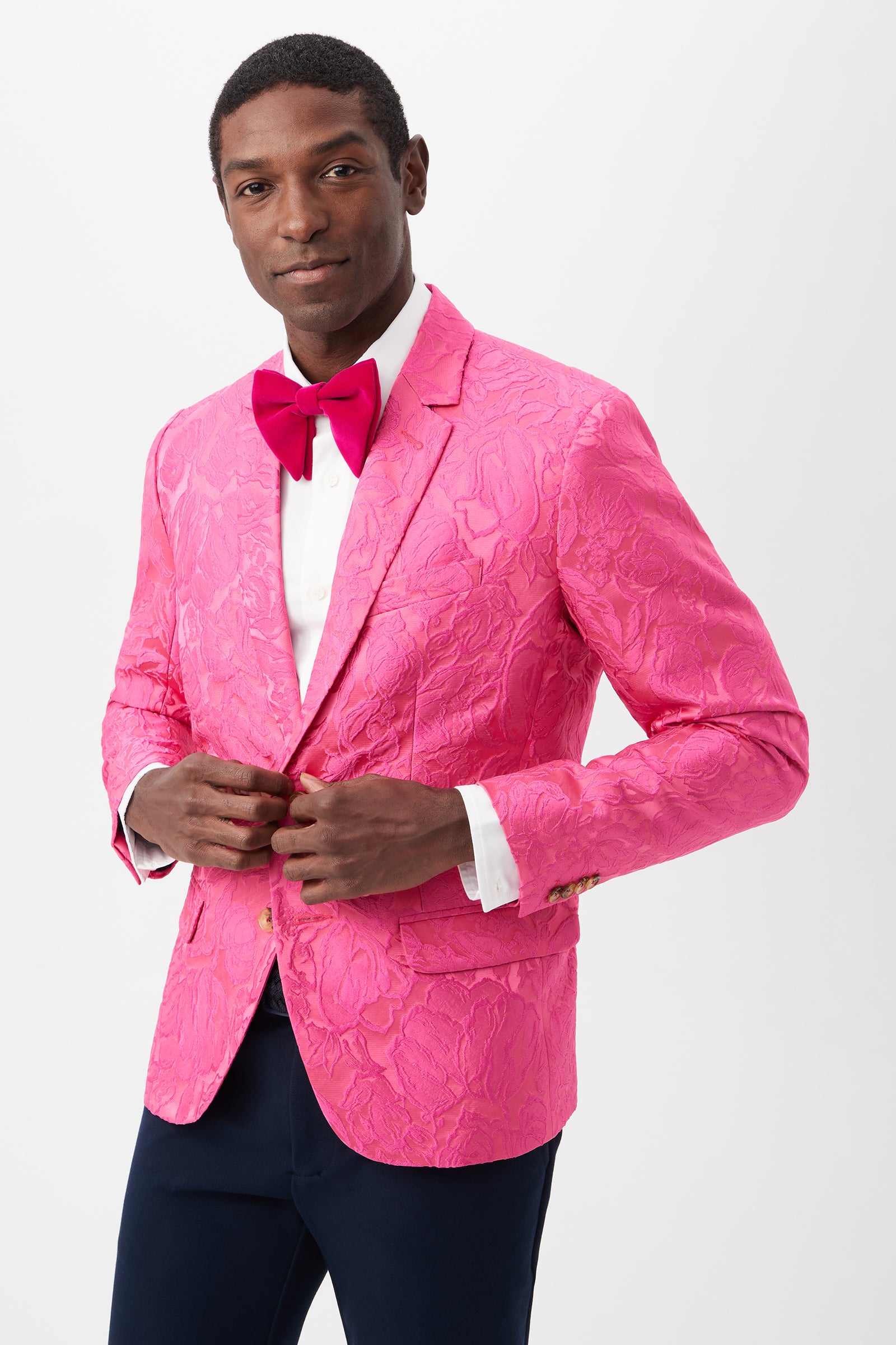 A man in the Thurston Blazer - Sweetheart Jacquard in Snapdragon, paired with a white dress shirt, pink bow tie, and dark pants, stands against a plain white background while buttoning his jacket and smiling slightly.