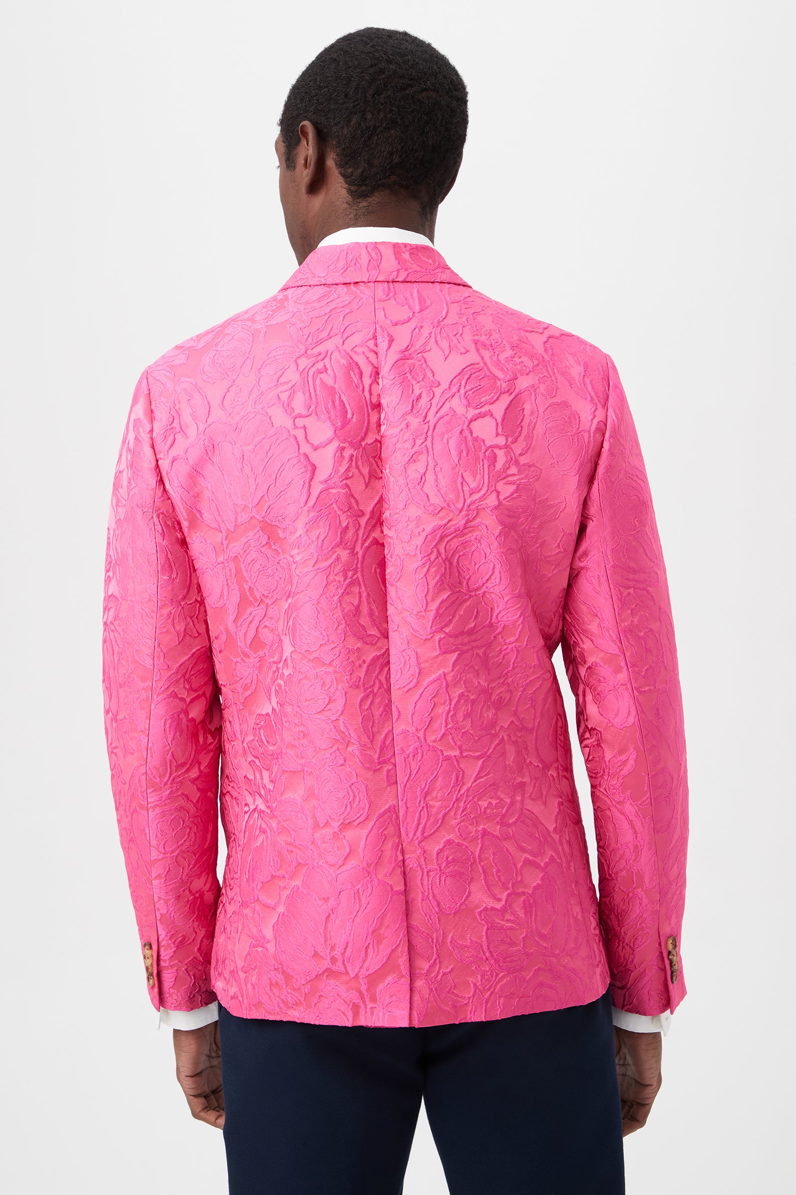 A person stands facing away in the Thurston Blazer - Sweetheart Jacquard, a bright pink textured floral-pattern blazer with visible white shirt cuffs and dark trousers, set against a plain white background.