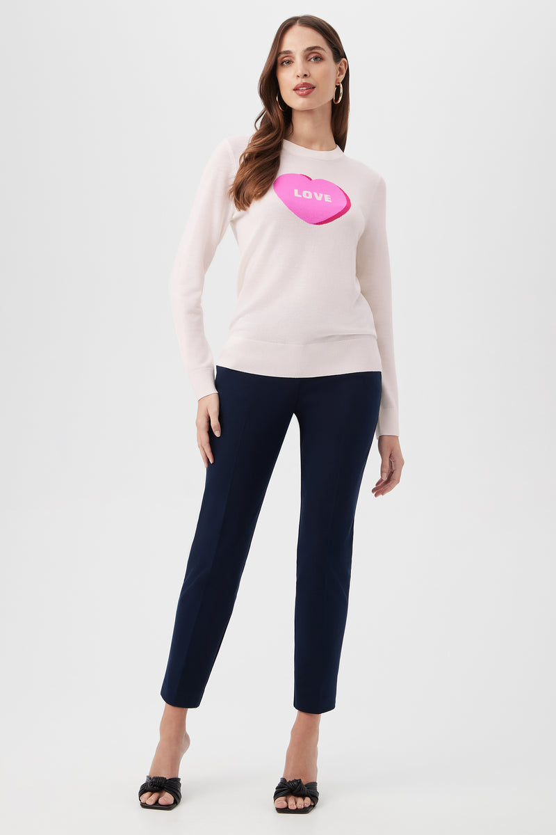 A woman wears the Love Merino Wool Crewneck Sweater in light pink with a large pink candy heart labeled LOVE, paired with navy ankle-length pants and black open-toe heels, standing against a plain white background.