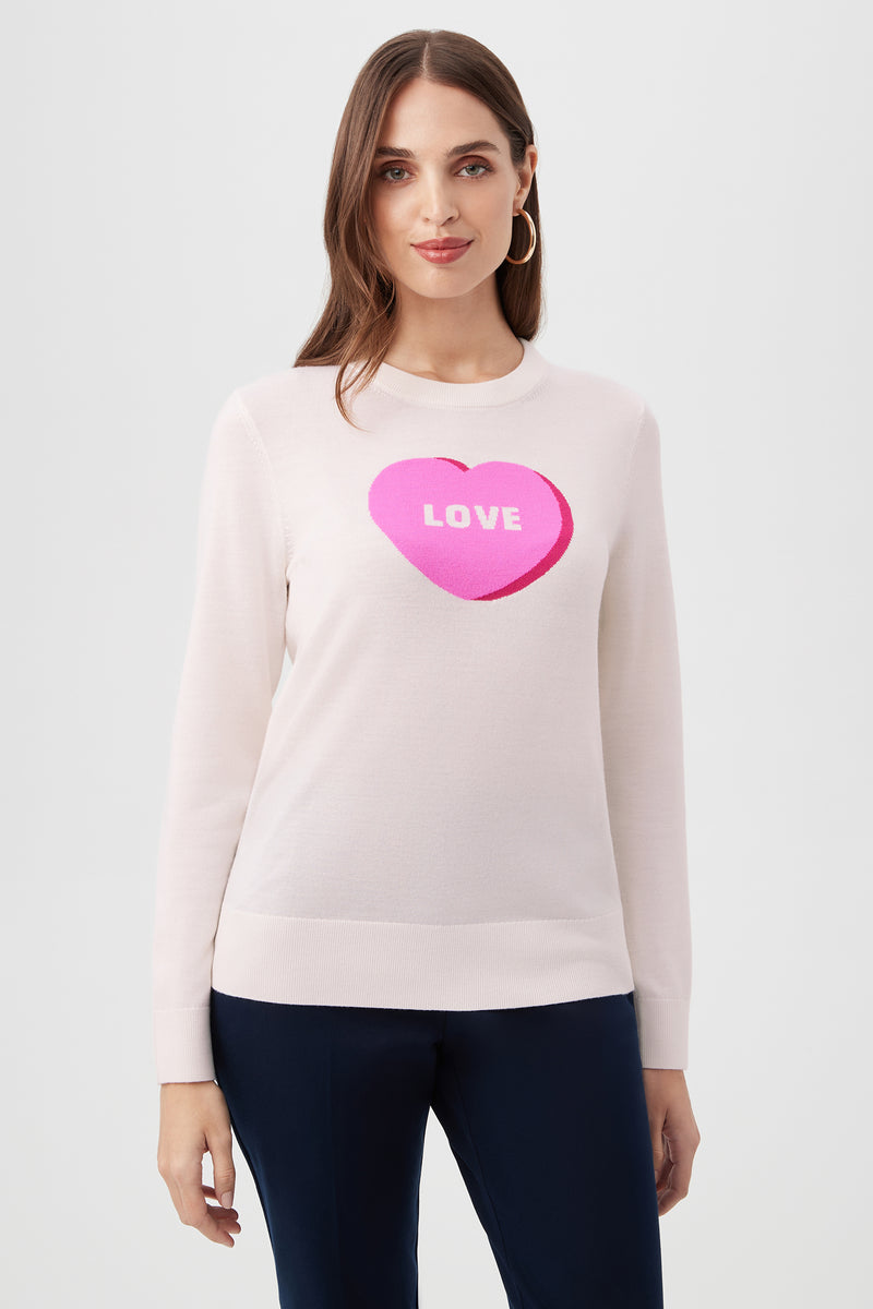 A woman with long brown hair wears the multi-colored Love Merino Wool Crewneck Sweater, which features a large pink heart with LOVE in white letters on the front, paired with dark pants, standing against a plain light background.