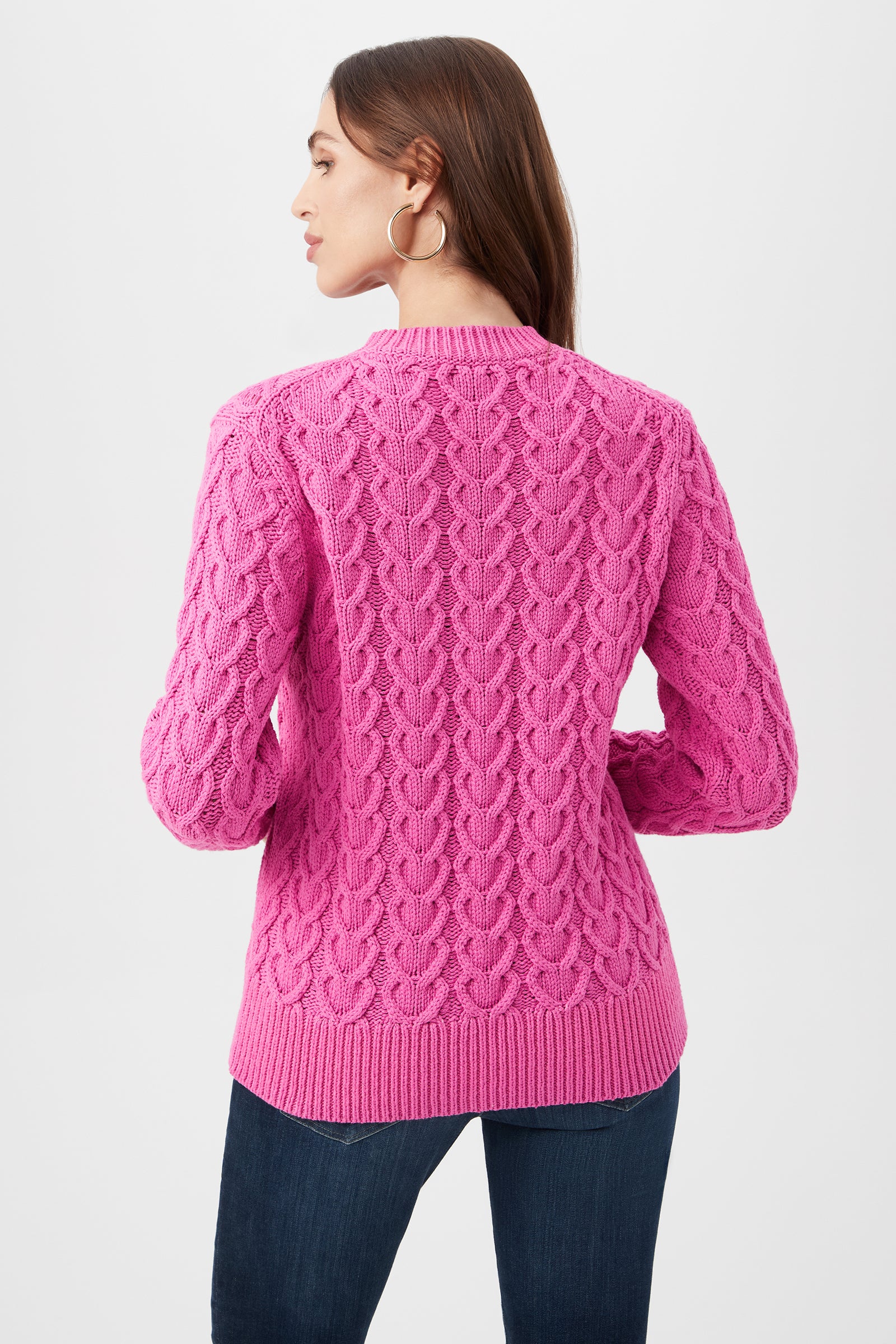A woman with long brown hair models the Corazon Heart Knit Crewneck Sweater in bright pink, paired with dark jeans. Facing away from the camera, she highlights the sweater’s textured back and wears large hoop earrings.