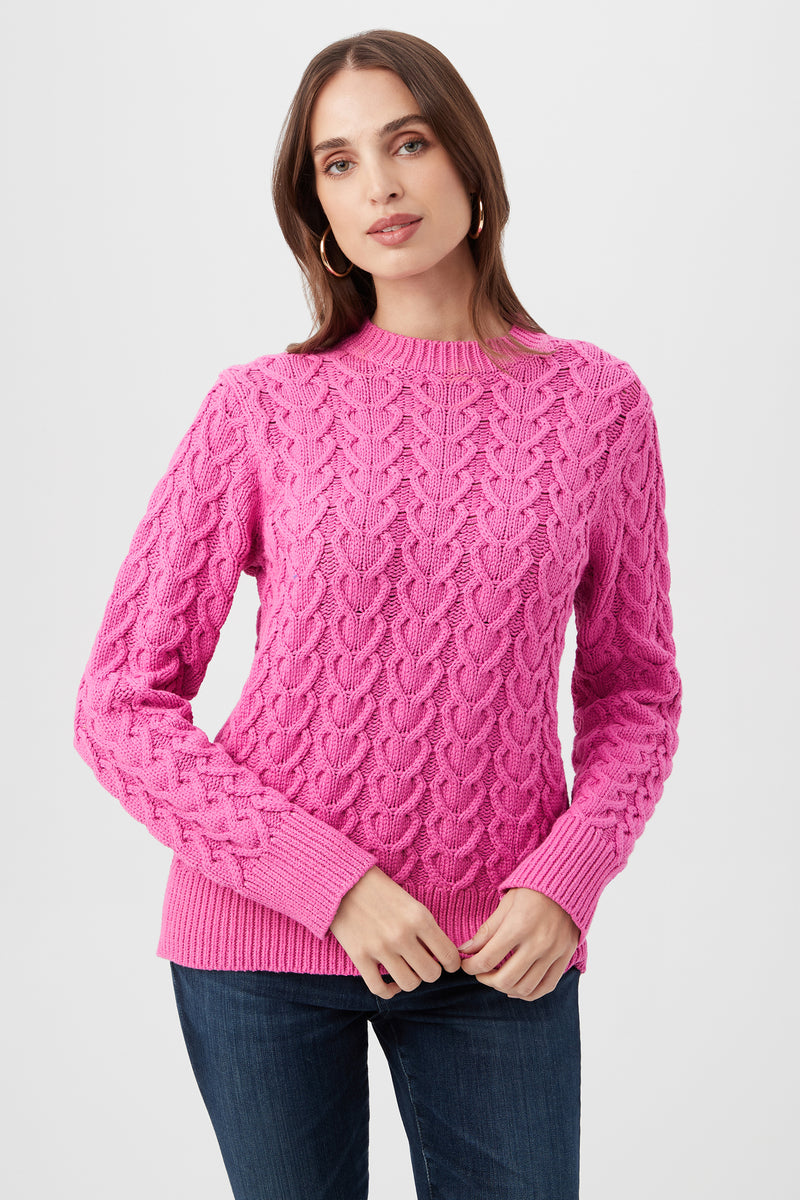 A woman wearing the Corazon Heart Knit Crewneck Sweater in Snapdragon and blue jeans stands against a plain light background, looking at the camera with a neutral expression.