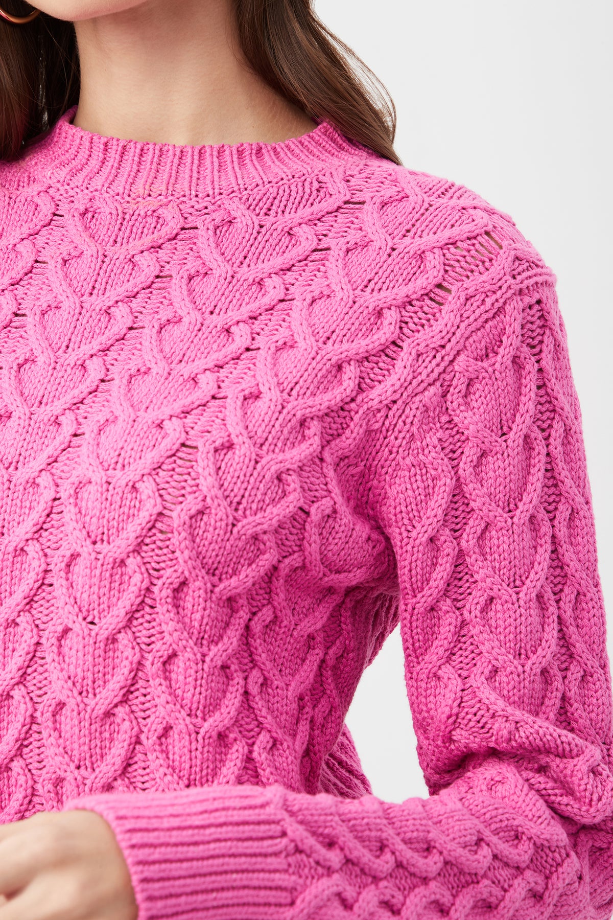 A close-up shows a person wearing the Corazon Heart Knit Crewneck Sweater in bright pink, featuring a textured cable-knit pattern. Only the lower face, neck, and upper torso are visible.