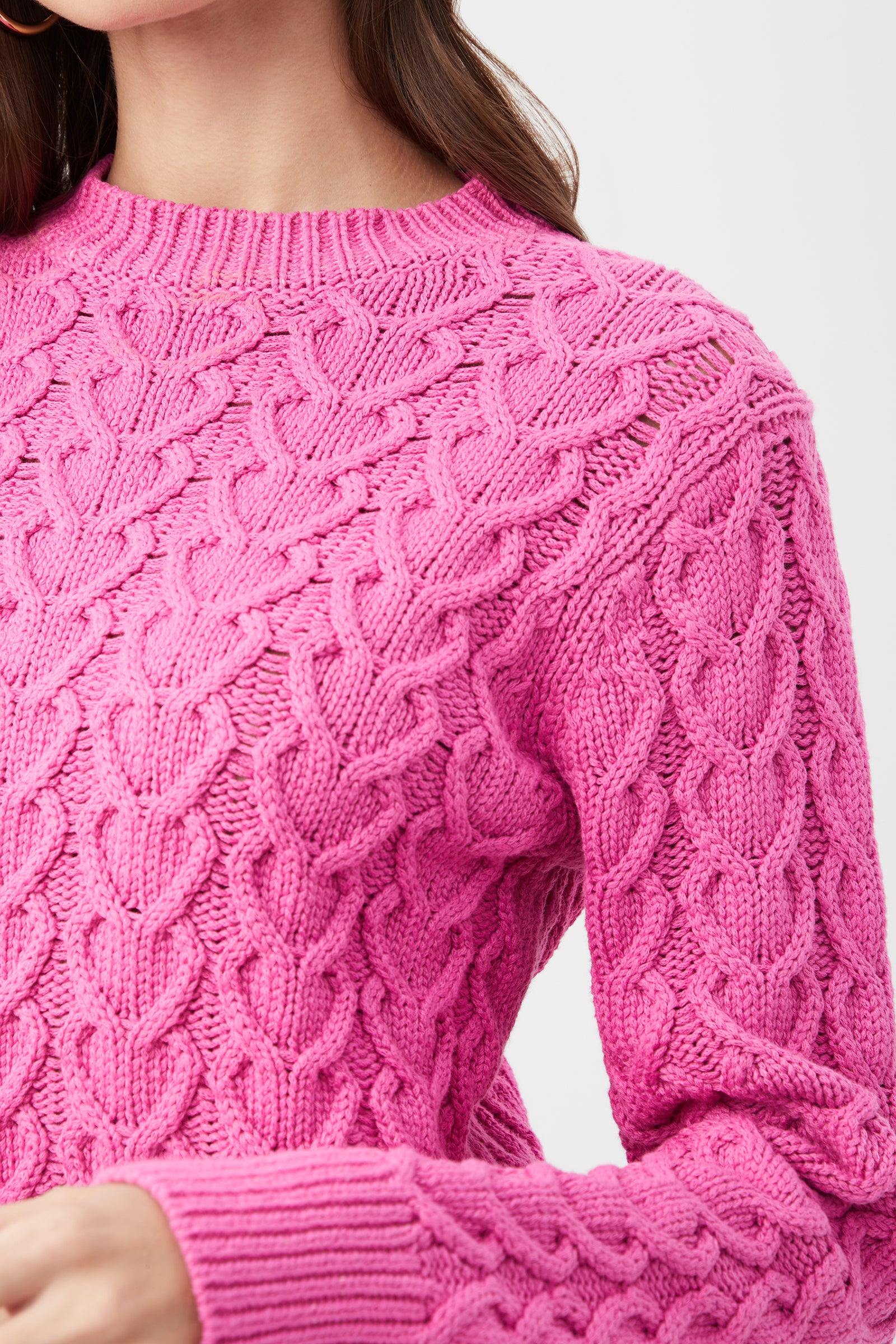 A close-up shows a person wearing the Corazon Heart Knit Crewneck Sweater in bright pink, featuring a textured cable-knit pattern. Only the lower face, neck, and upper torso are visible.