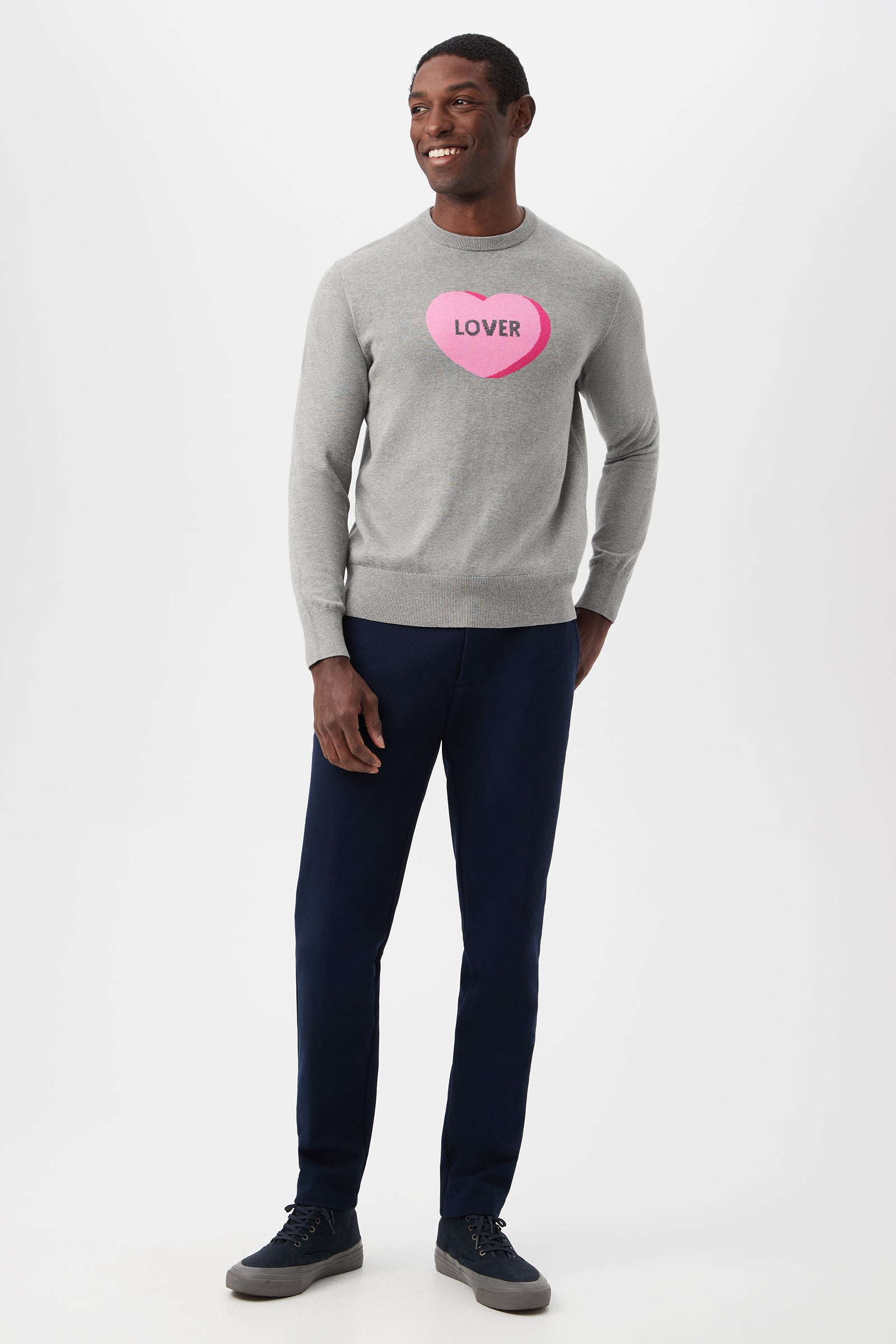 A man stands smiling in the Inamorato Sweetheart Crewneck Sweater—a light gray sweater with a pink heart and LOVER on it—paired with dark blue pants and dark sneakers, set against a plain white background.