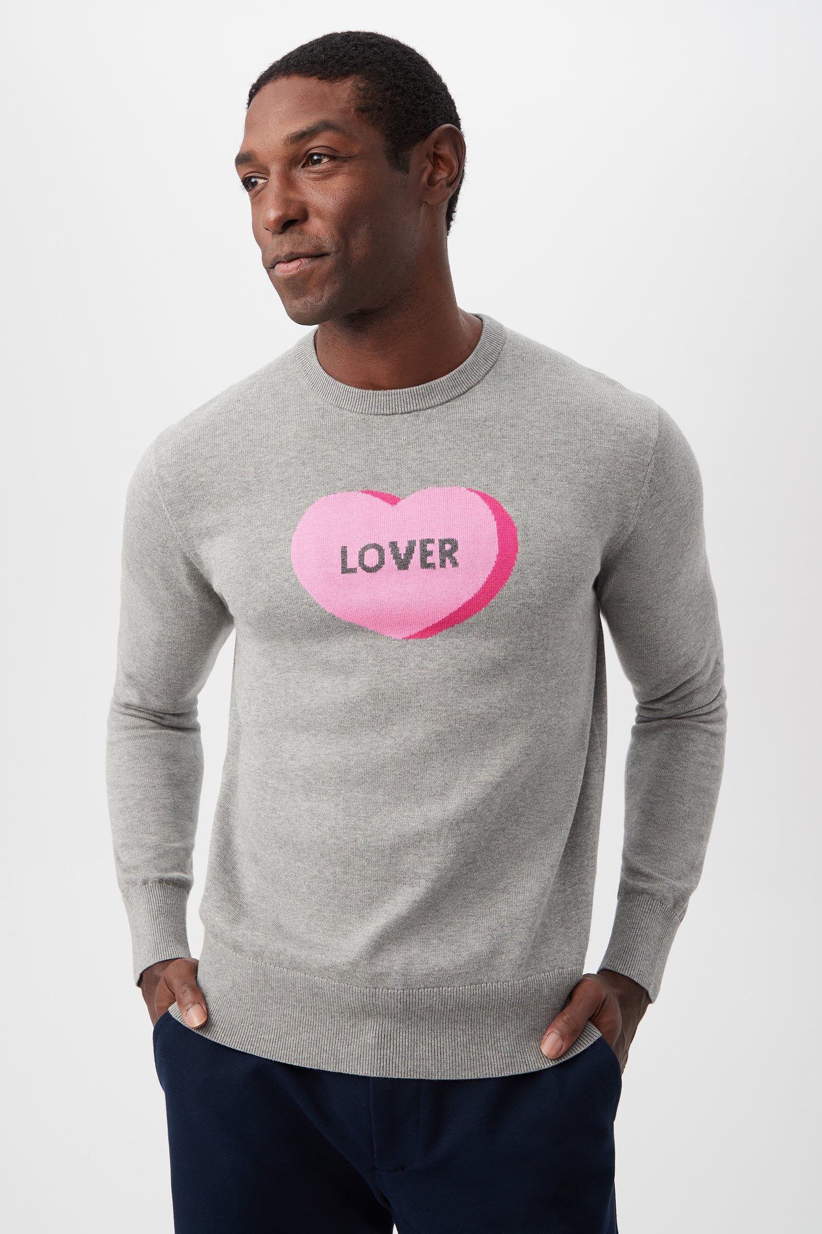 A man stands with hands in pockets, wearing the Inamorato Sweetheart Crewneck Sweater in heather grey with a large pink heart and LOVER on the chest, set against a plain light background.