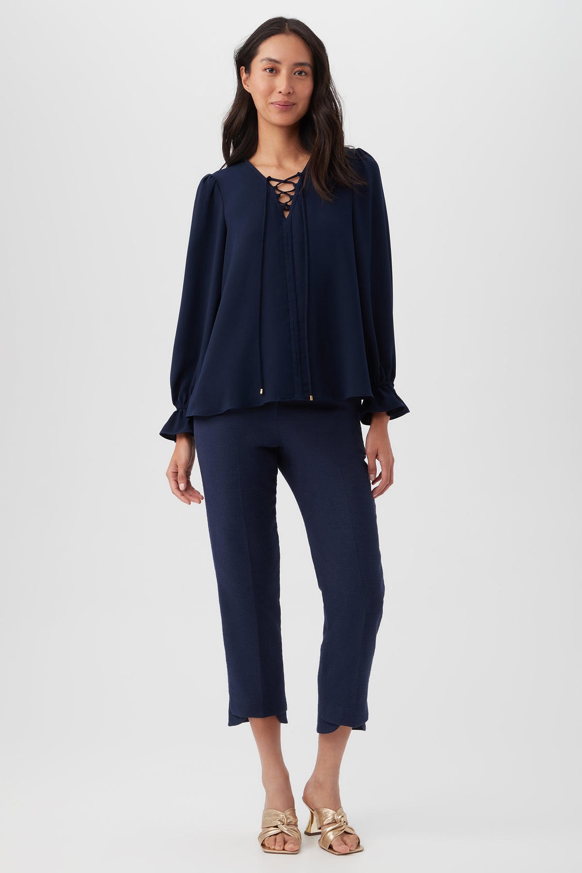 A woman stands against a plain background wearing the Zahara Top, a loose satin-back crepe blouse with long balloon sleeves, paired with cropped navy pants and gold flat sandals.