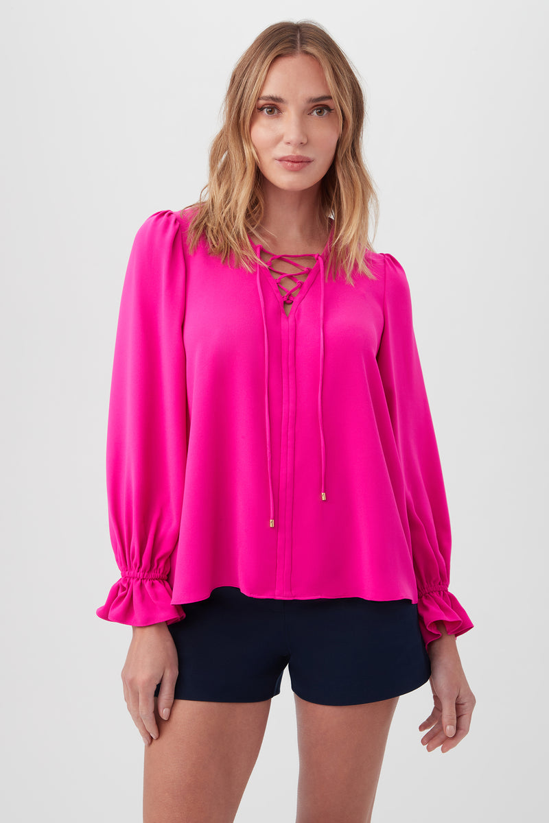 A woman with shoulder-length blonde hair wears the Zahara Top in Trina Pink, featuring balloon sleeves and a lace-up neckline, paired with dark shorts against a plain light background.