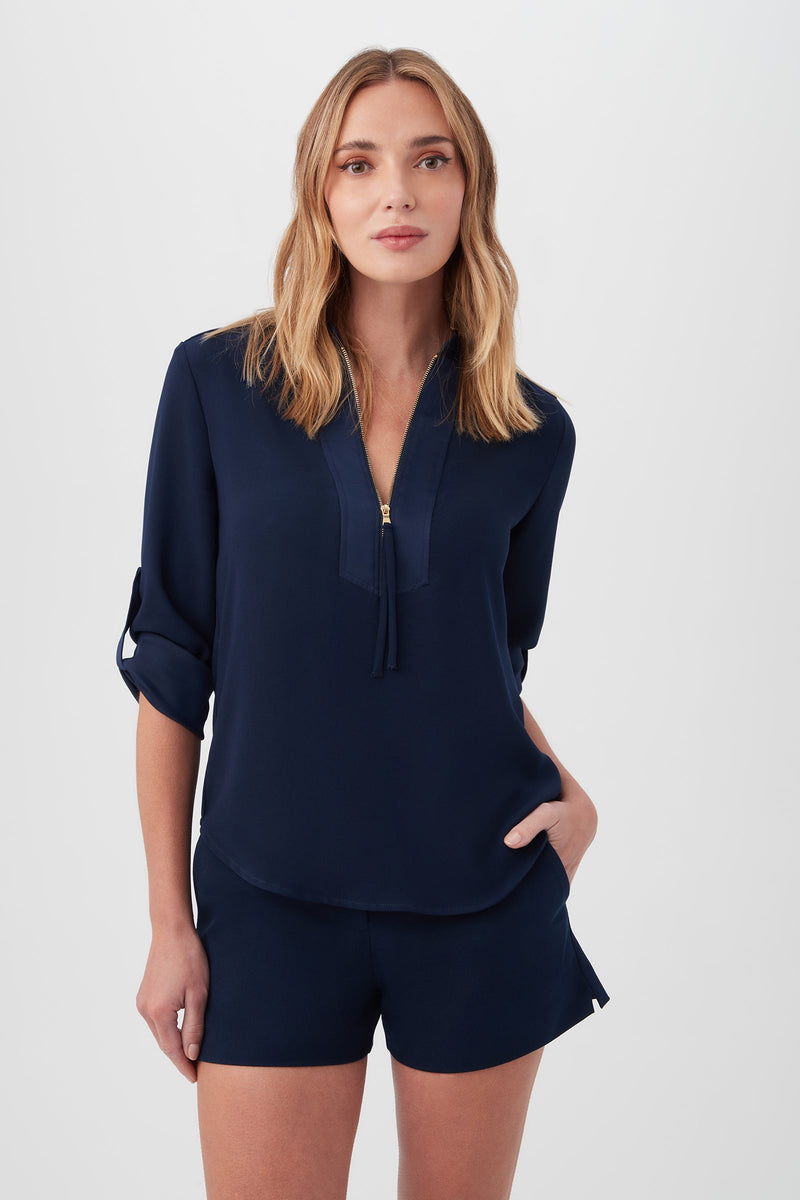 A woman with light brown hair wears the Kaiko 3/4-Sleeve Half-Zip Top in indigo, made from sustainable fabric, standing against a plain light background with one hand in her pocket.