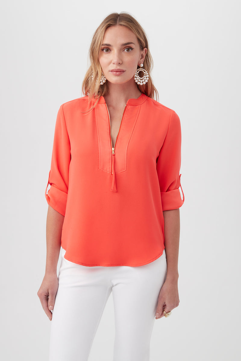 A woman wears the Kaiko 3/4-Sleeve Half-Zip Top in bright coral Silky Satin-Back Crepe, featuring a collarless V-neck and tassel detail, paired with white pants. She has wavy blonde hair and statement earrings against a plain background.