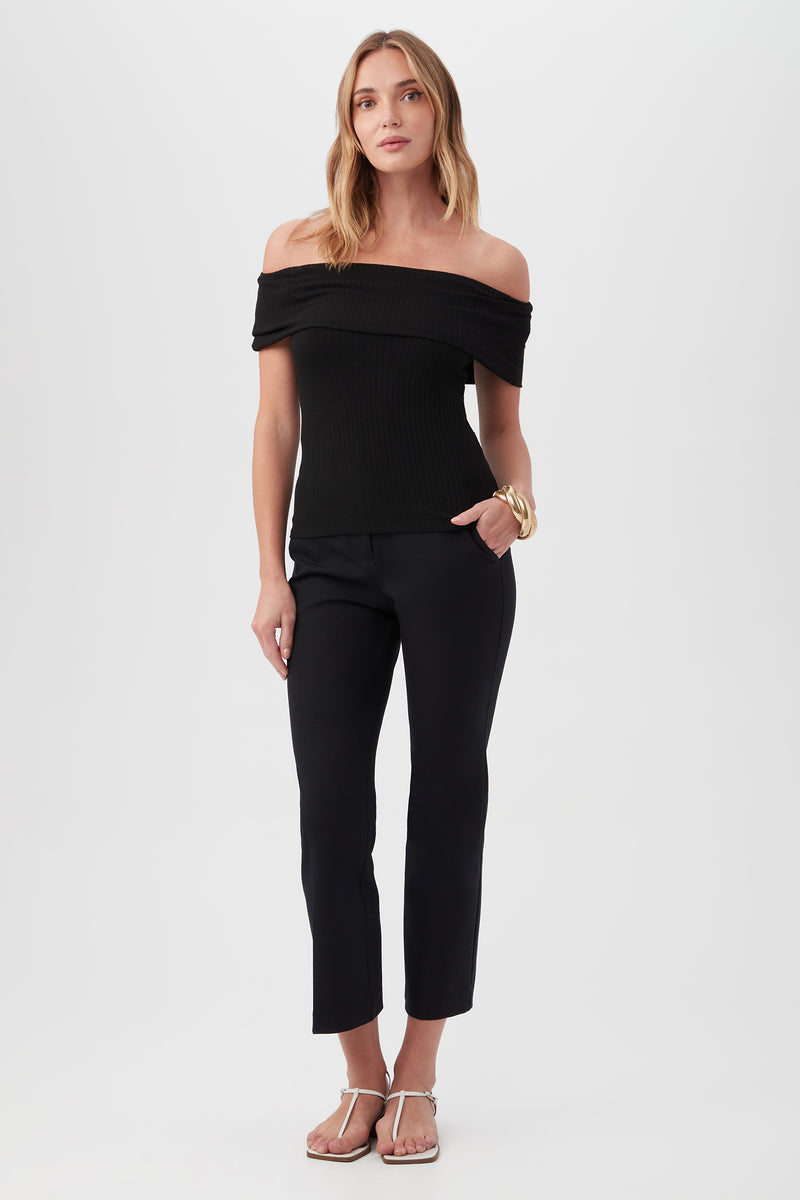A woman poses against a white background, wearing the Prizma Off-Shoulder Top in black with black cropped pants, white sandals, and gold bracelets. She has one hand in her pocket and a relaxed expression.