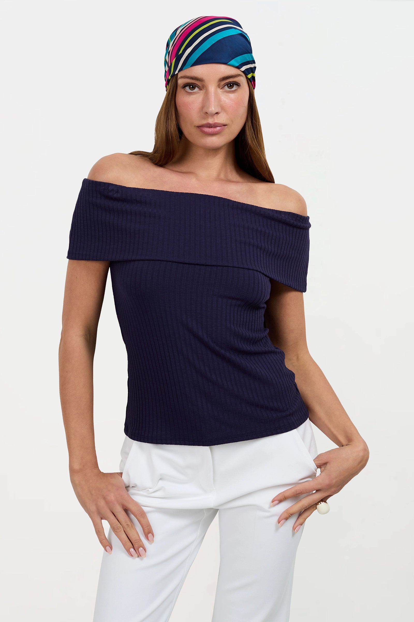 A woman wears the Prizma Off-Shoulder Top with a colorful headscarf and white pants, posing with one hand on her hip against a plain light background.