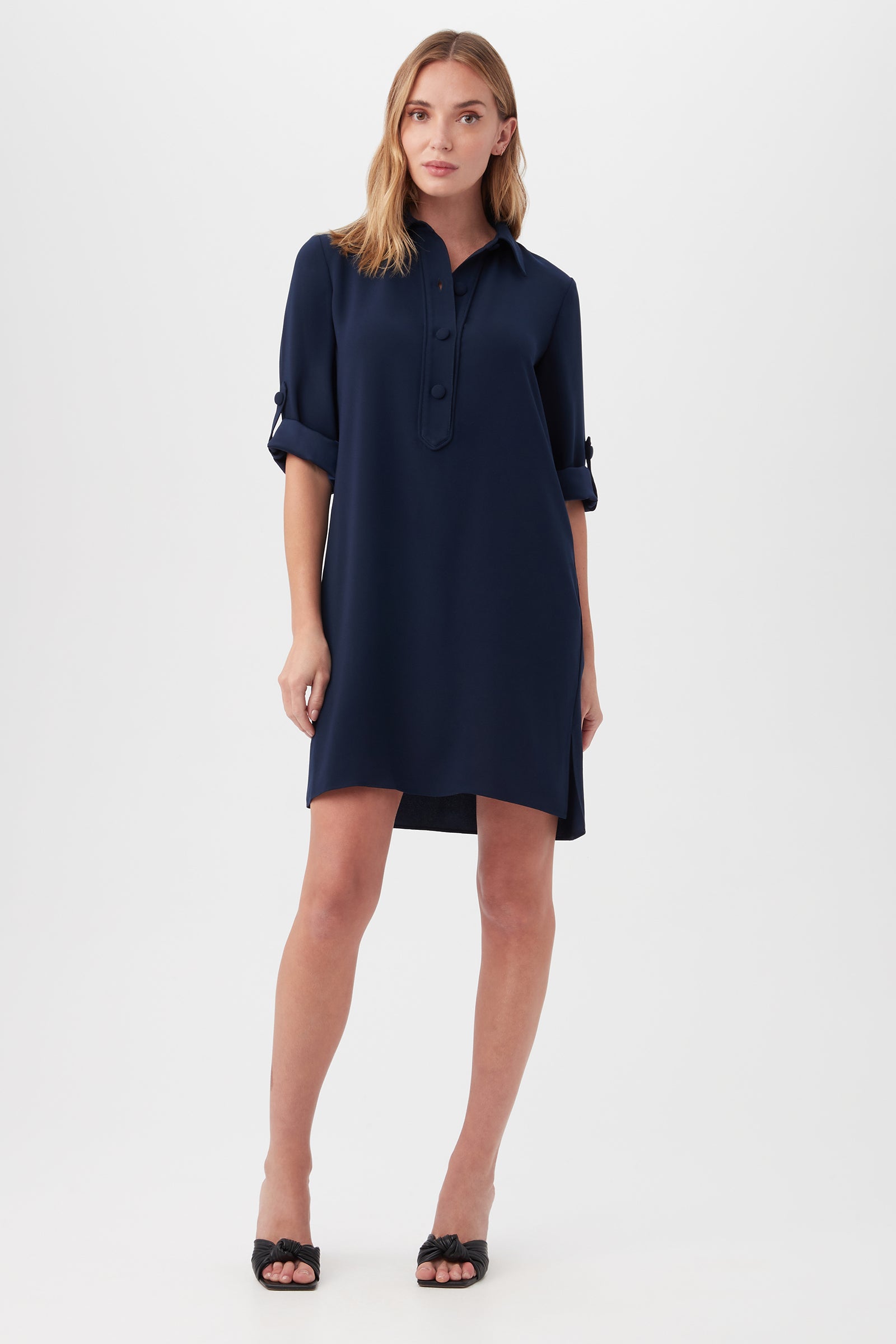 A woman with long, light brown hair stands against a white background wearing the Portrait Shirt Dress in indigo with buttons, a collar, rolled-up sleeves, and knee-length hem. She completes the look with black open-toe heels.