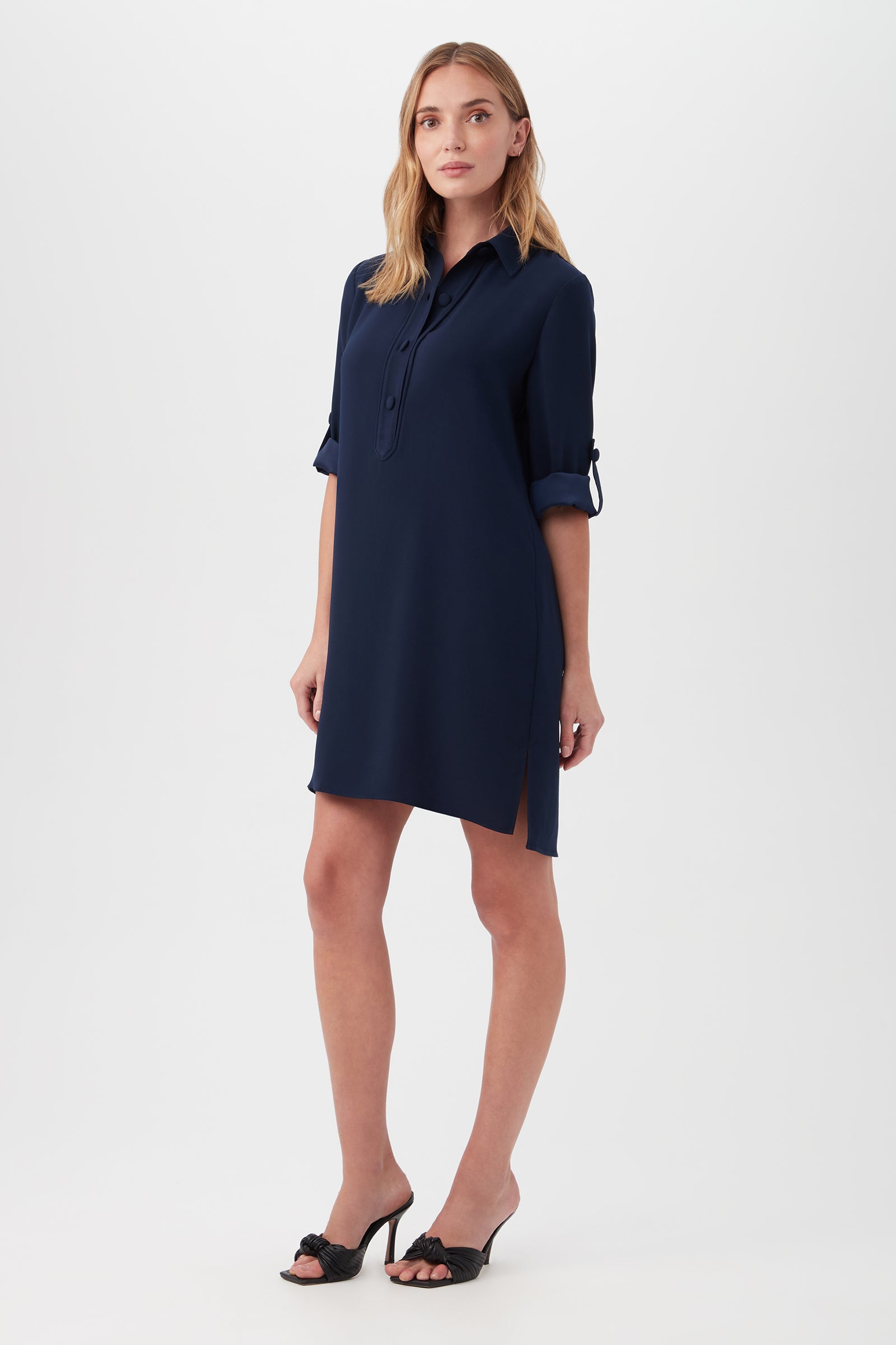 A woman wears the Portrait Shirt Dress in navy blue, featuring a knee-length cut, rolled-up sleeves, and a collar. She pairs it with black heeled sandals and poses against a plain white background.