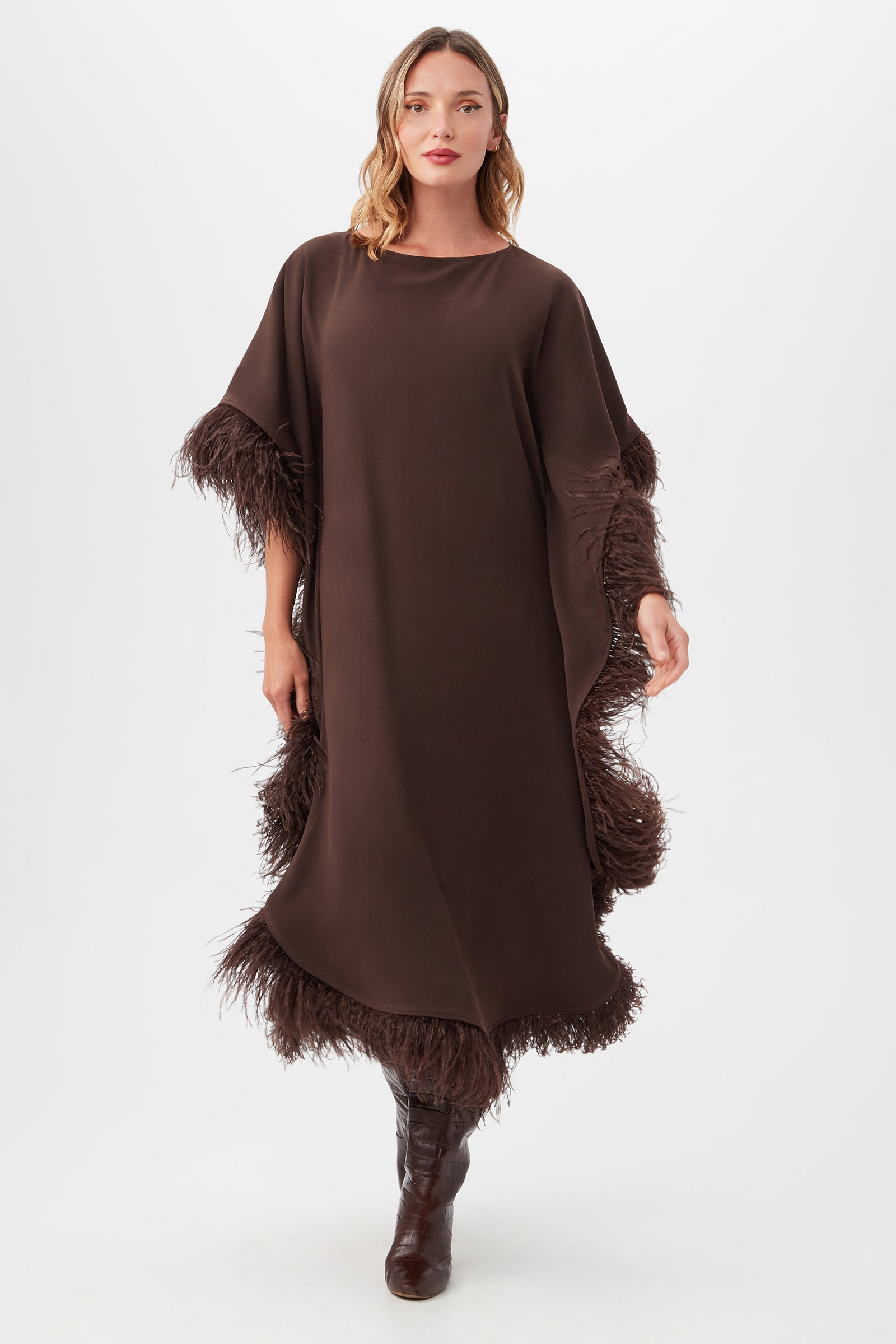 A woman models the Neena Maxi Caftan Dress in Brown Derby, featuring feathered trim on the sleeves and hem. She pairs it with dark brown boots and poses against a plain white background.