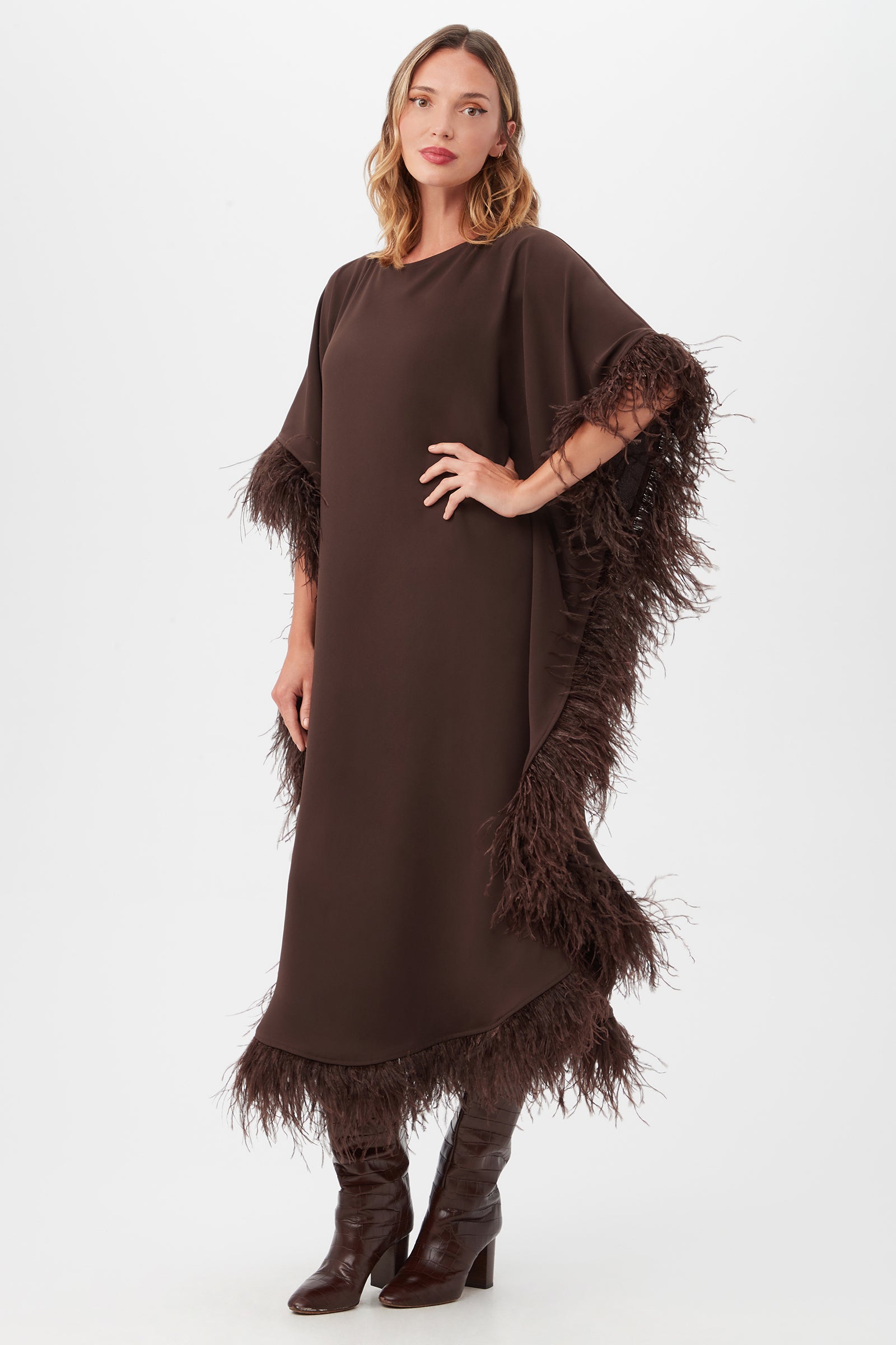 A woman models the Neena Maxi Caftan Dress, a loose dark brown dress with feathery trim on the sleeves and hem, styled with matching knee-high boots against a plain white background.