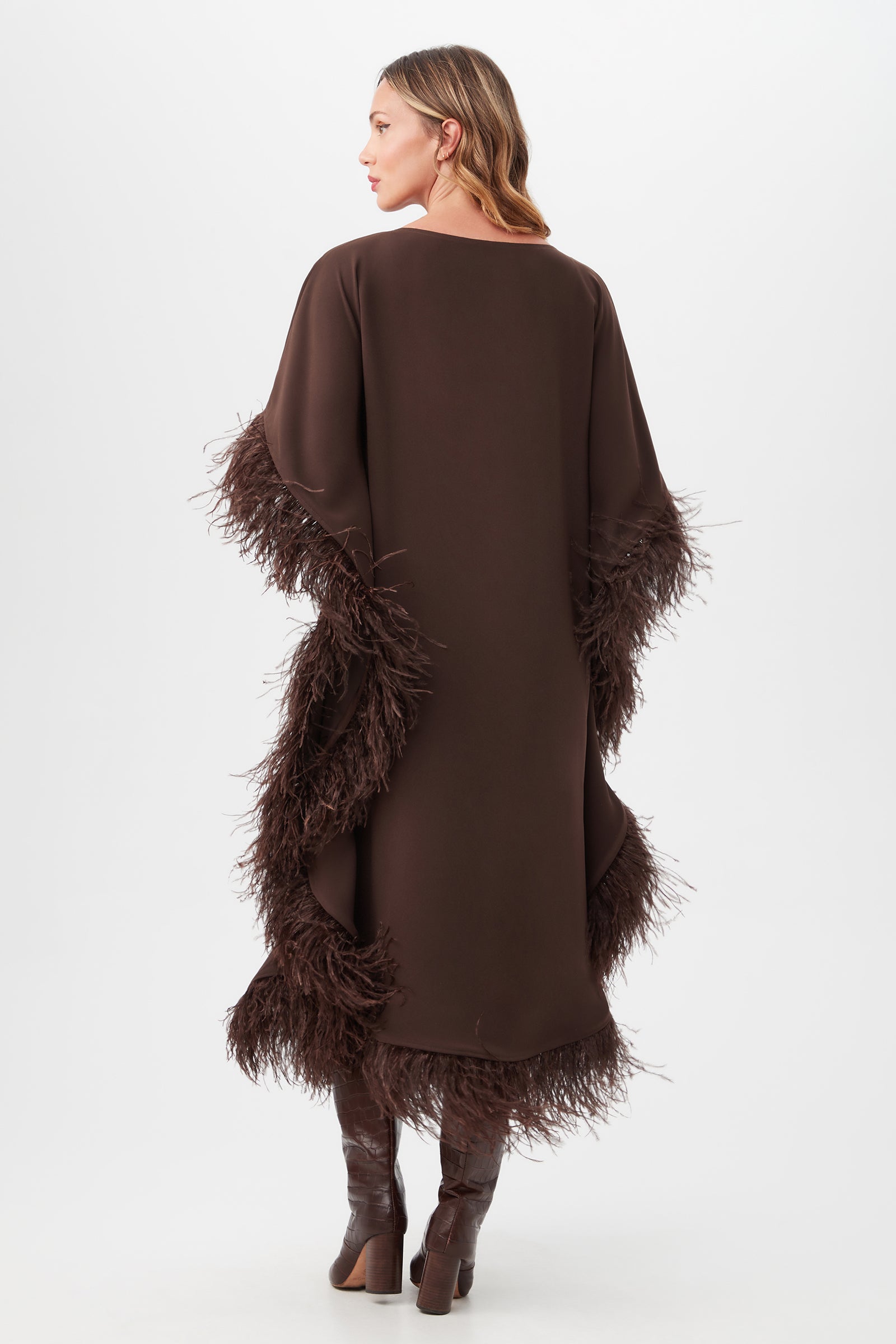 A woman with wavy hair faces away, wearing the Neena Maxi Caftan Dress in dark brown with feathered trim on the sides and sleeves, paired with tall brown boots, set against a plain white background.