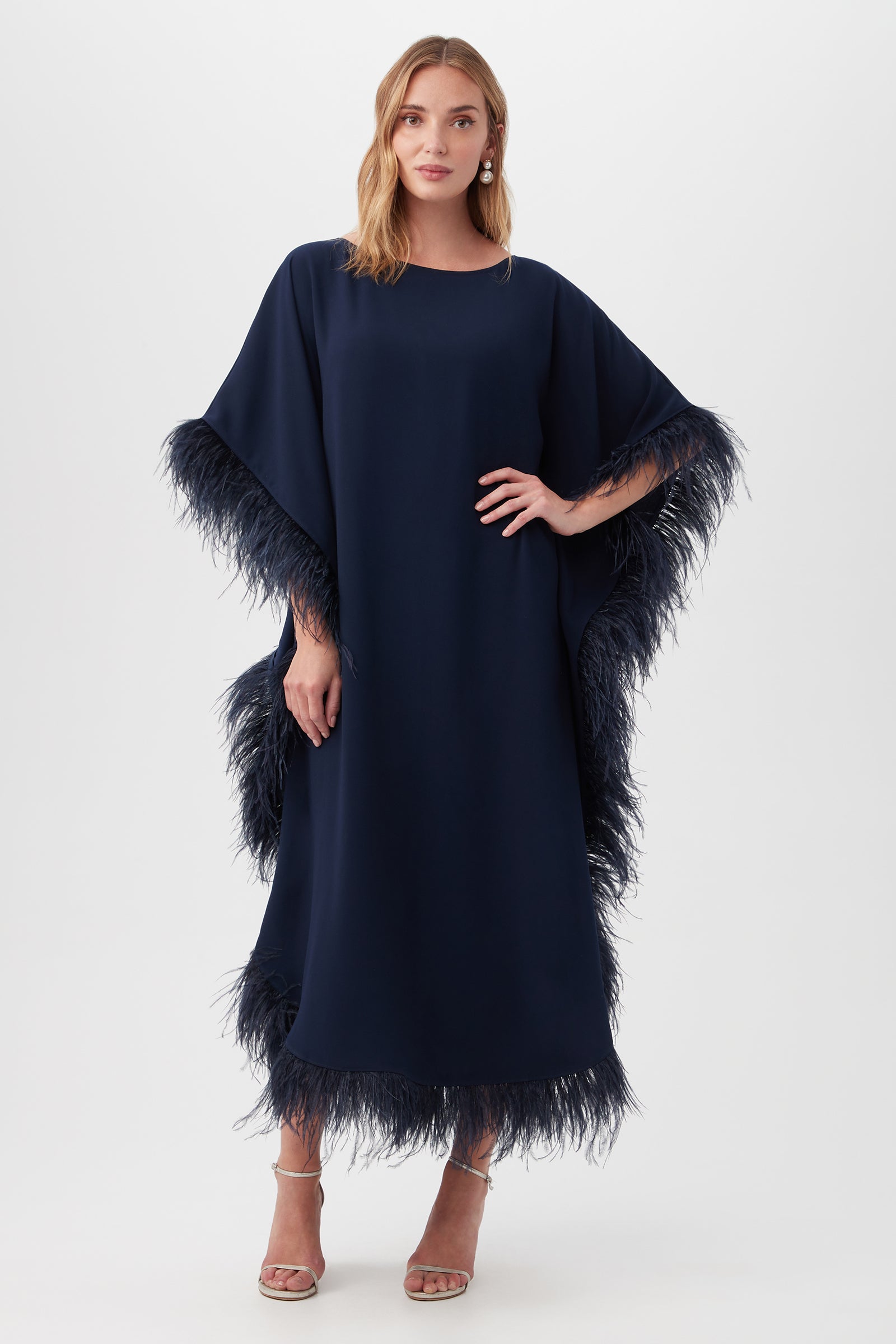 A woman with light brown hair stands facing forward in the Neena Maxi Caftan Dress in indigo, featuring feathery trim on the sleeves and hem. She wears open-toed heels, set against a plain white background.