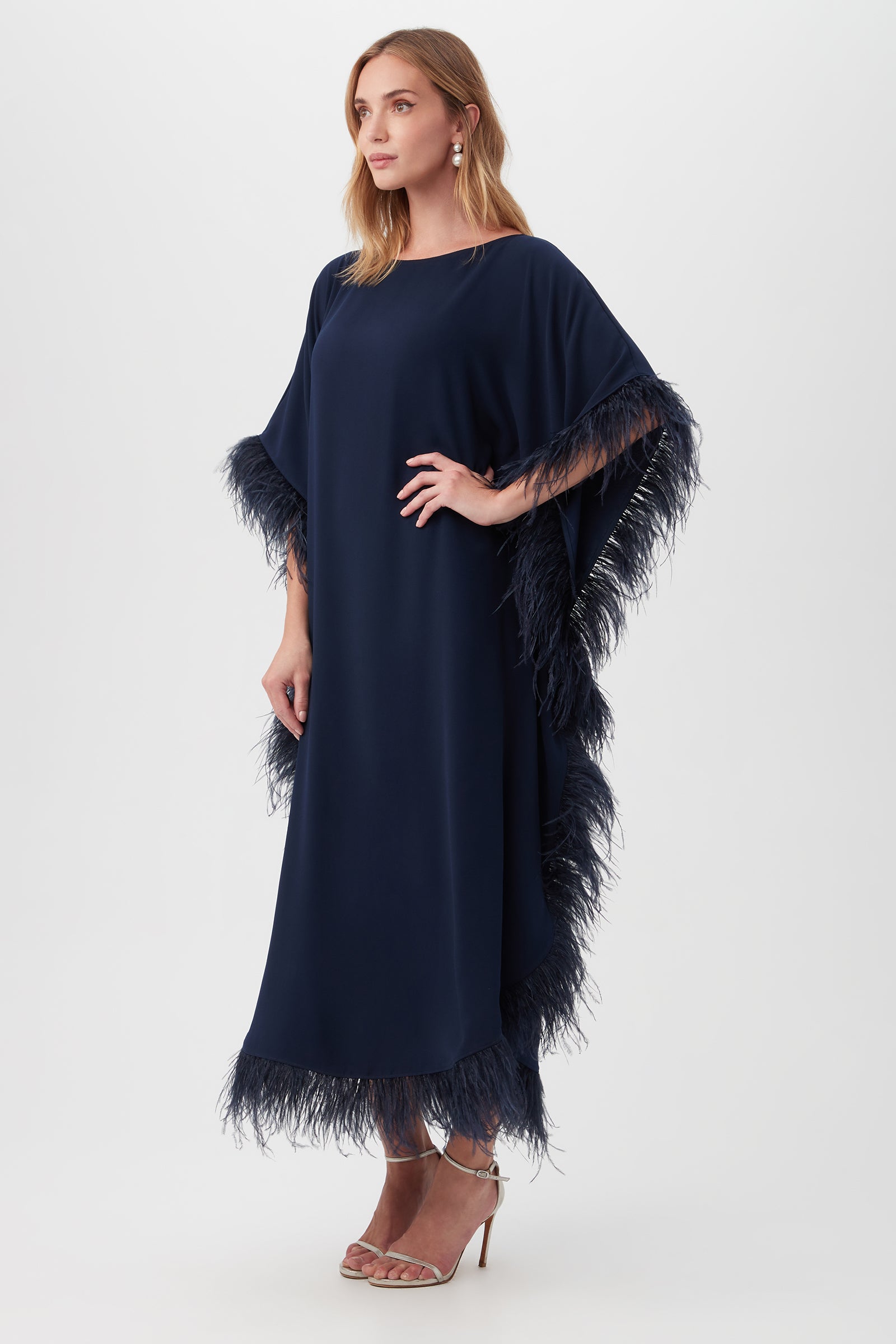 A woman models the Neena Maxi Caftan Dress—a loose-fitting navy blue style with feather-trimmed sleeves and hem. She has light brown hair, earrings, and nude heels, posing against a plain white backdrop.