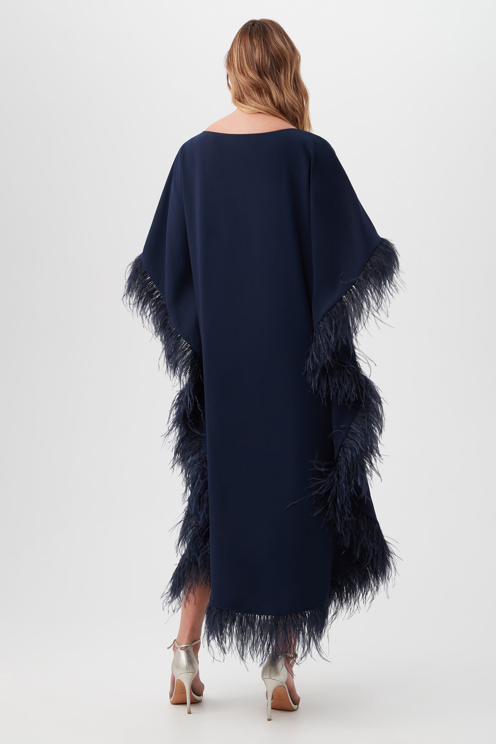 A woman with light brown wavy hair, in silver heels, faces away while wearing the Neena Maxi Caftan Dress— a long, loose-fitting navy blue dress with feather trim on the sleeves and hem—against a plain white background.