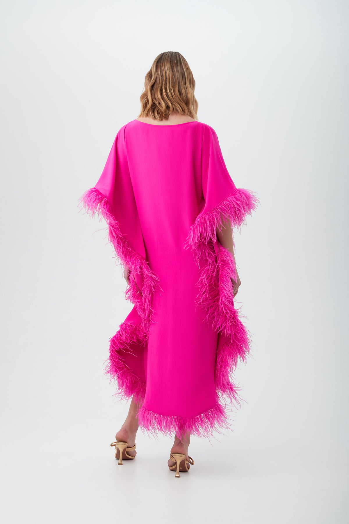 A woman with wavy, shoulder-length hair stands facing away, wearing the bright pink Neena Maxi Caftan Dress featuring feather trim on the sleeves and sides, paired with tan high-heeled sandals.