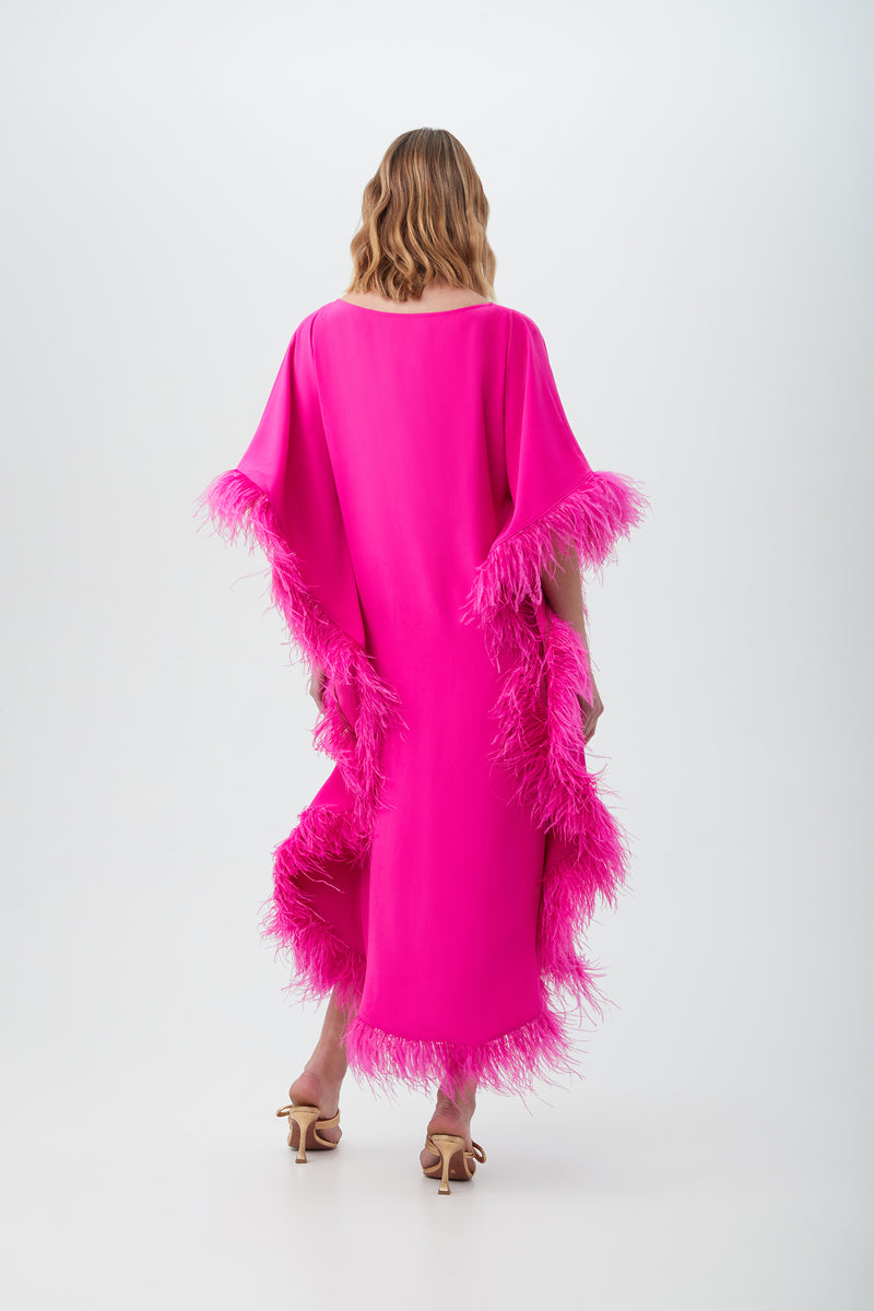 A woman with wavy, shoulder-length hair stands facing away, wearing the bright pink Neena Maxi Caftan Dress featuring feather trim on the sleeves and sides, paired with tan high-heeled sandals.