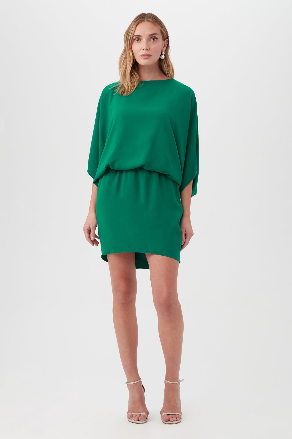 A woman faces forward in the Manhattan Dress in emerald, featuring batwing sleeves and a cinched waist, paired with beige strappy high-heeled sandals against a plain white background.