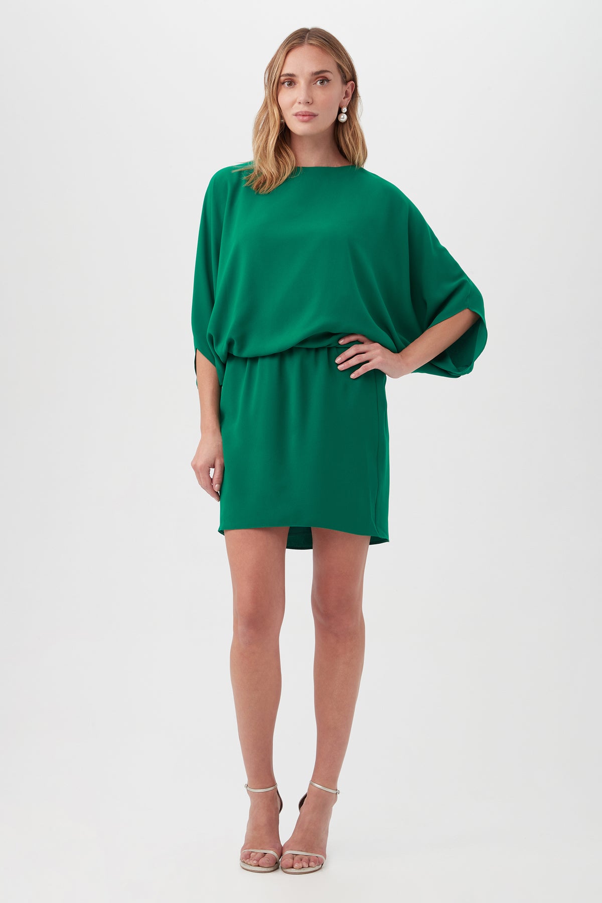 Woman stands against a white background wearing the Manhattan Dress—a loose, knee-length green dress with batwing sleeves and belted waist—paired with nude strappy high-heeled sandals.